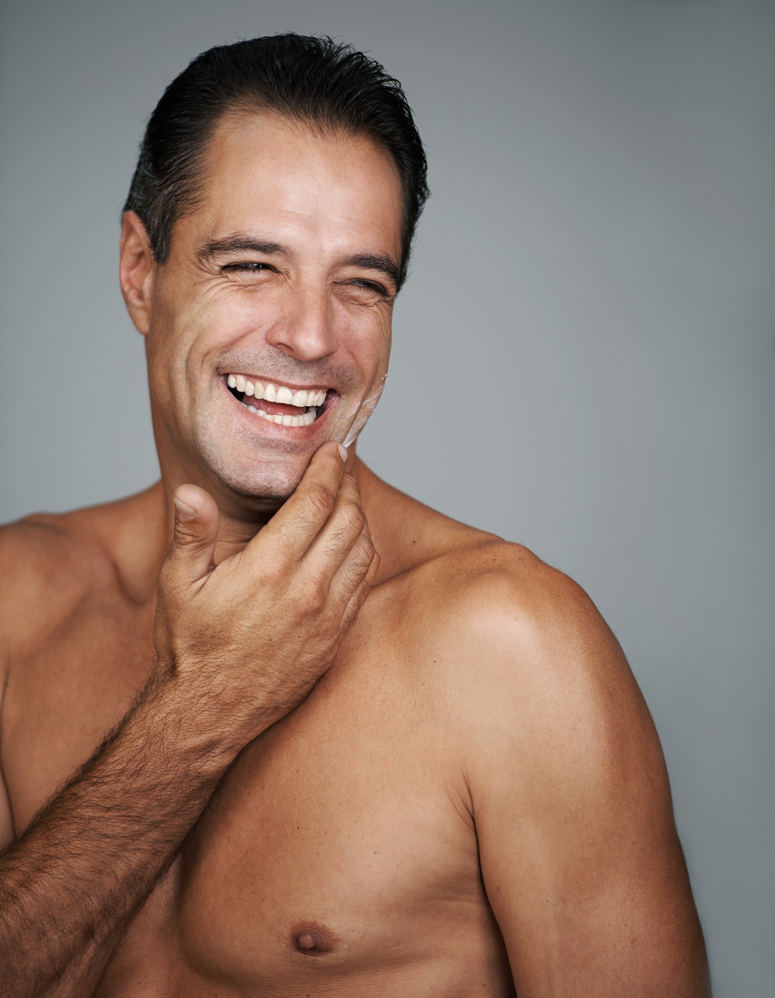 A shirtless man with dark hair and a broad smile touching his face with his right hand, standing against a plain gray background.