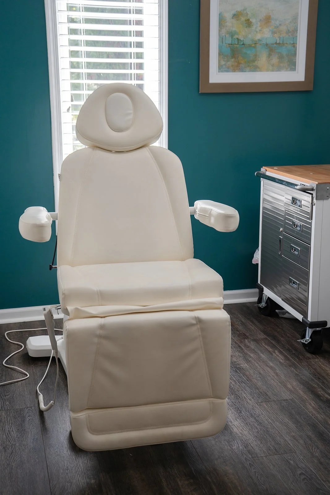 Medical examination chair in a clinic room with teal walls, a window with white blinds, and a framed abstract painting.