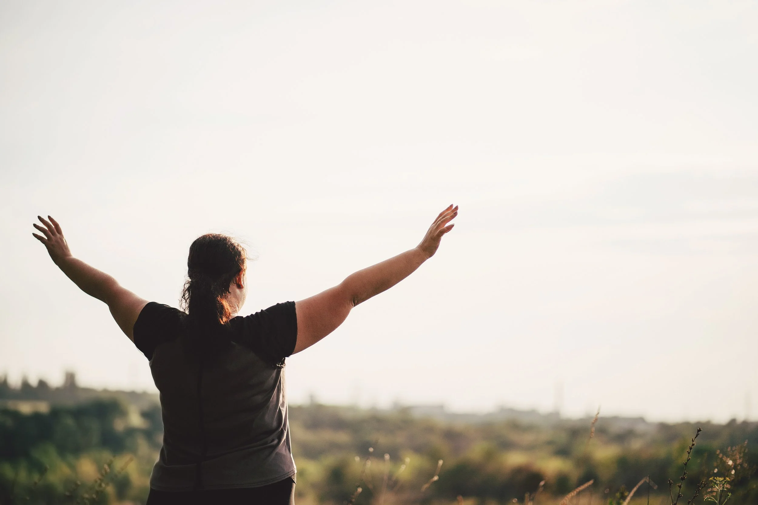 A person with dark hair and wearing a black shirt standing outdoors with arms raised, facing a bright sky with a blurred landscape in the background.