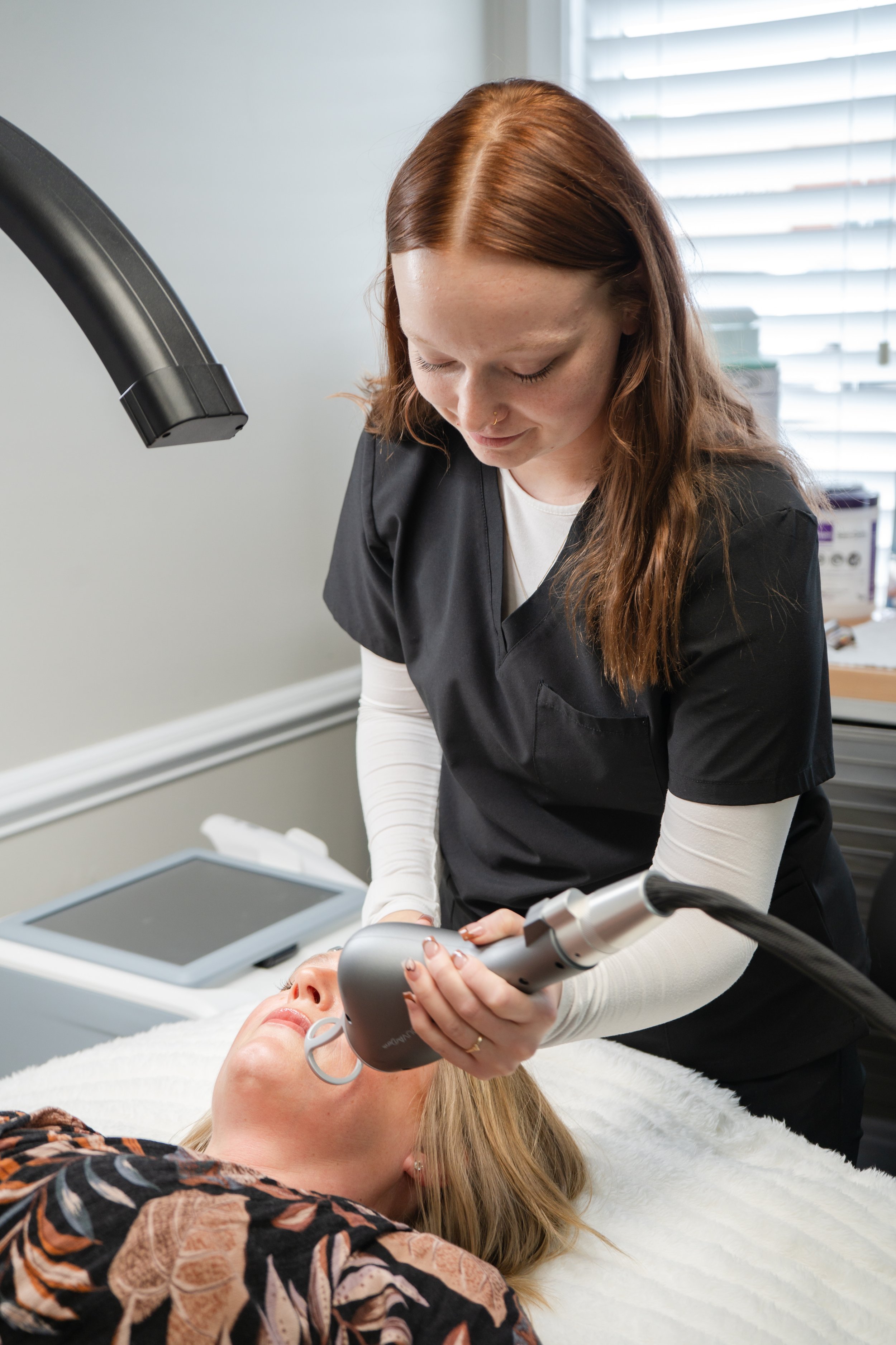 A woman with red hair performs a skin treatment using a handheld device on a woman lying on a table in a clinical setting.