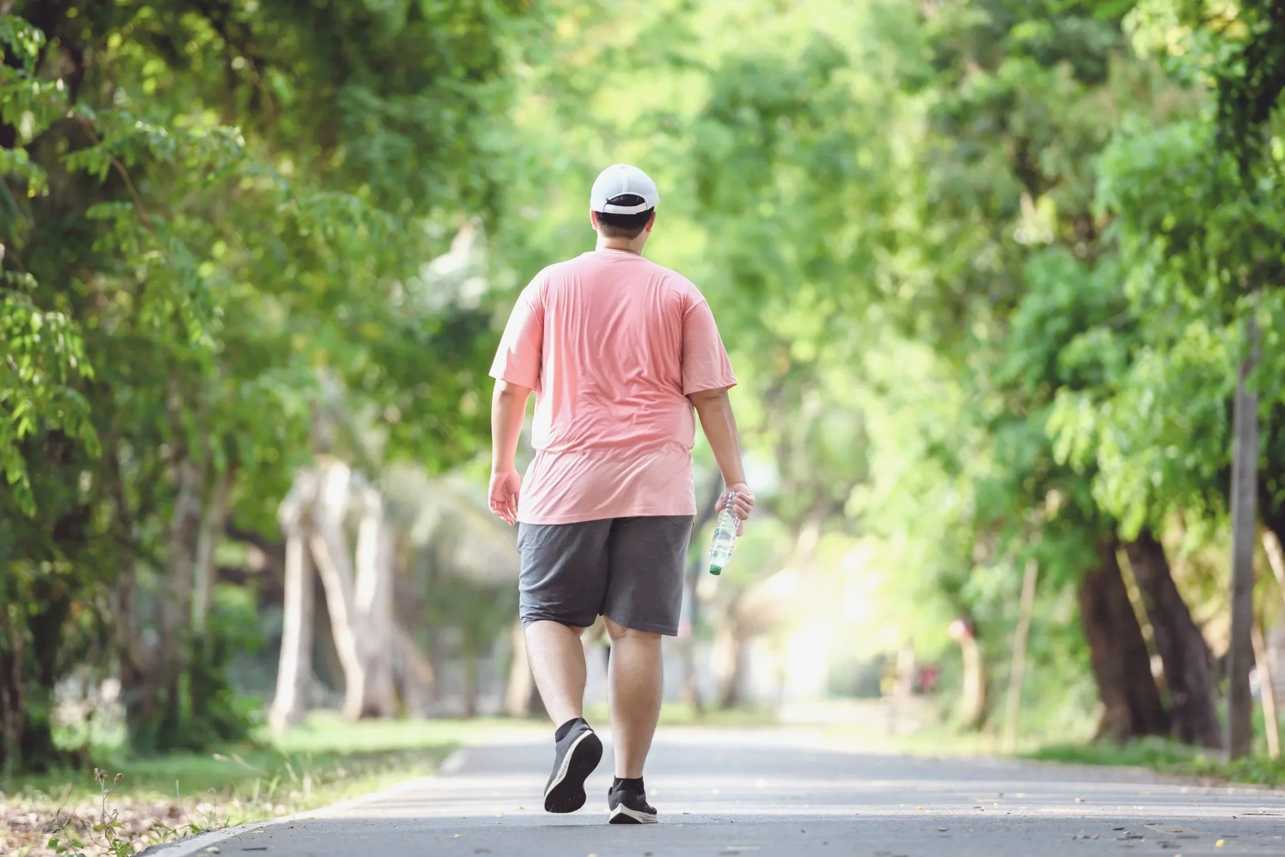 A man wearing a pink t-shirt, gray shorts, and a white baseball cap walking on a paved path through a green park, holding a water bottle in his right hand.