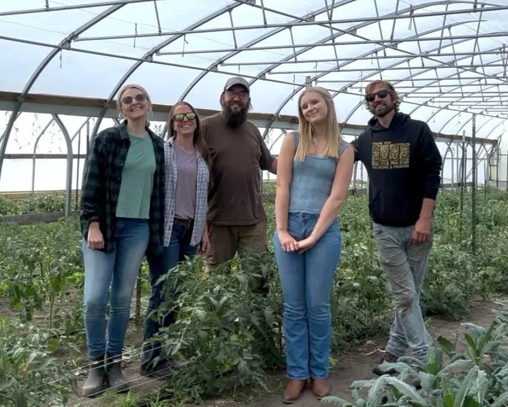 Five people standing inside a greenhouse, surrounded by green plants, smiling for the photo.