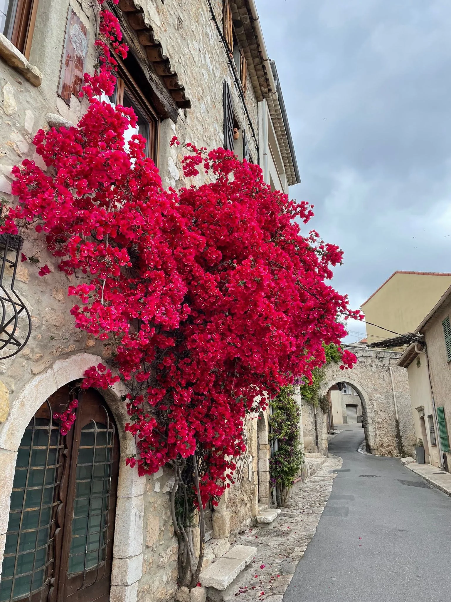 A stone building with pink bougainvillea climbing the wall next to arched windows and a small door. The street is narrow and winding with an archway at the end.