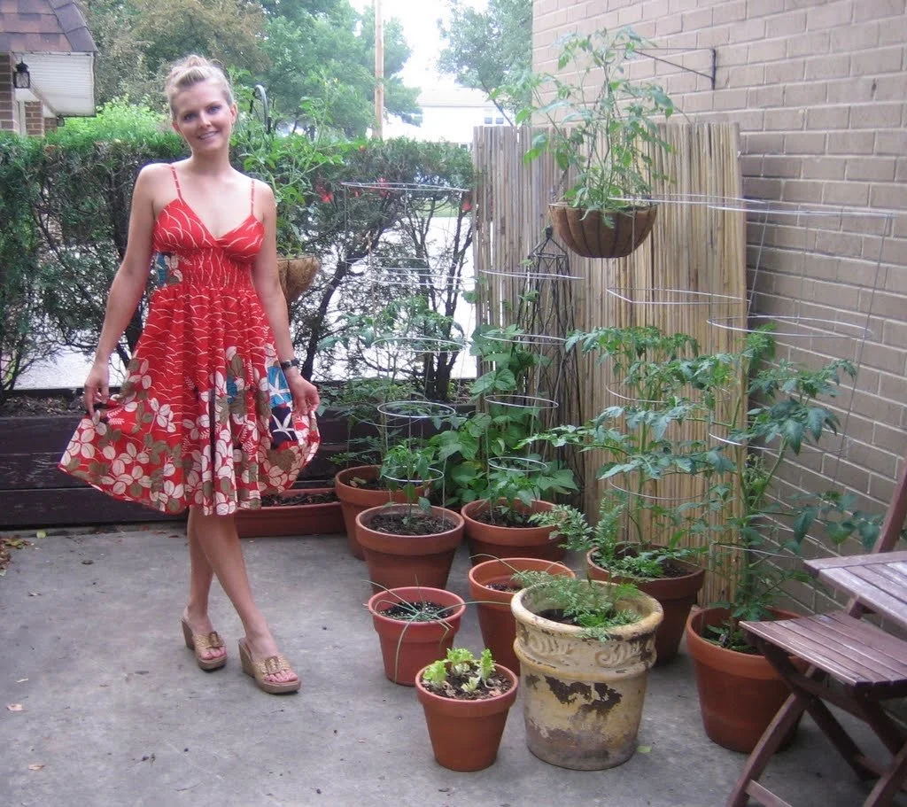 A young woman in a red floral dress and platform shoes standing next to a variety of potted plants on a patio, with a wooden privacy screen and shrubs in the background.