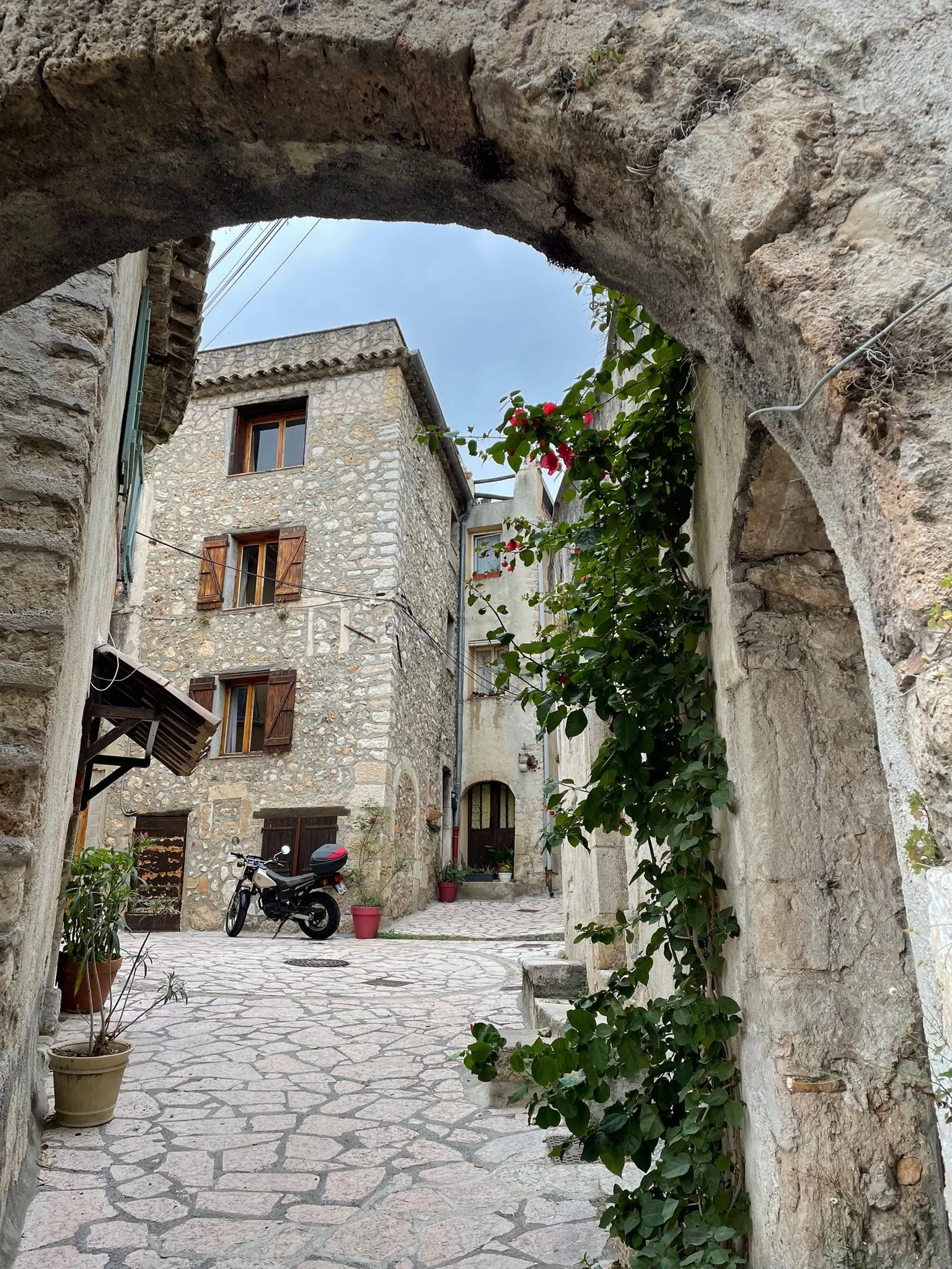 A rustic stone alleyway with an archway framing historic buildings, potted plants, and a parked motorcycle.