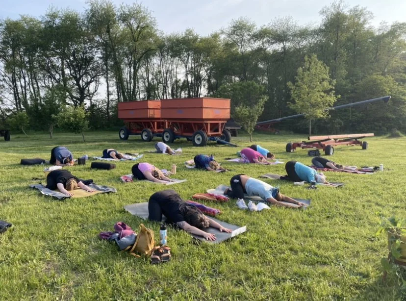 People participating in an outdoor yoga or meditation session on a grassy field, with trees and farm equipment in the background.