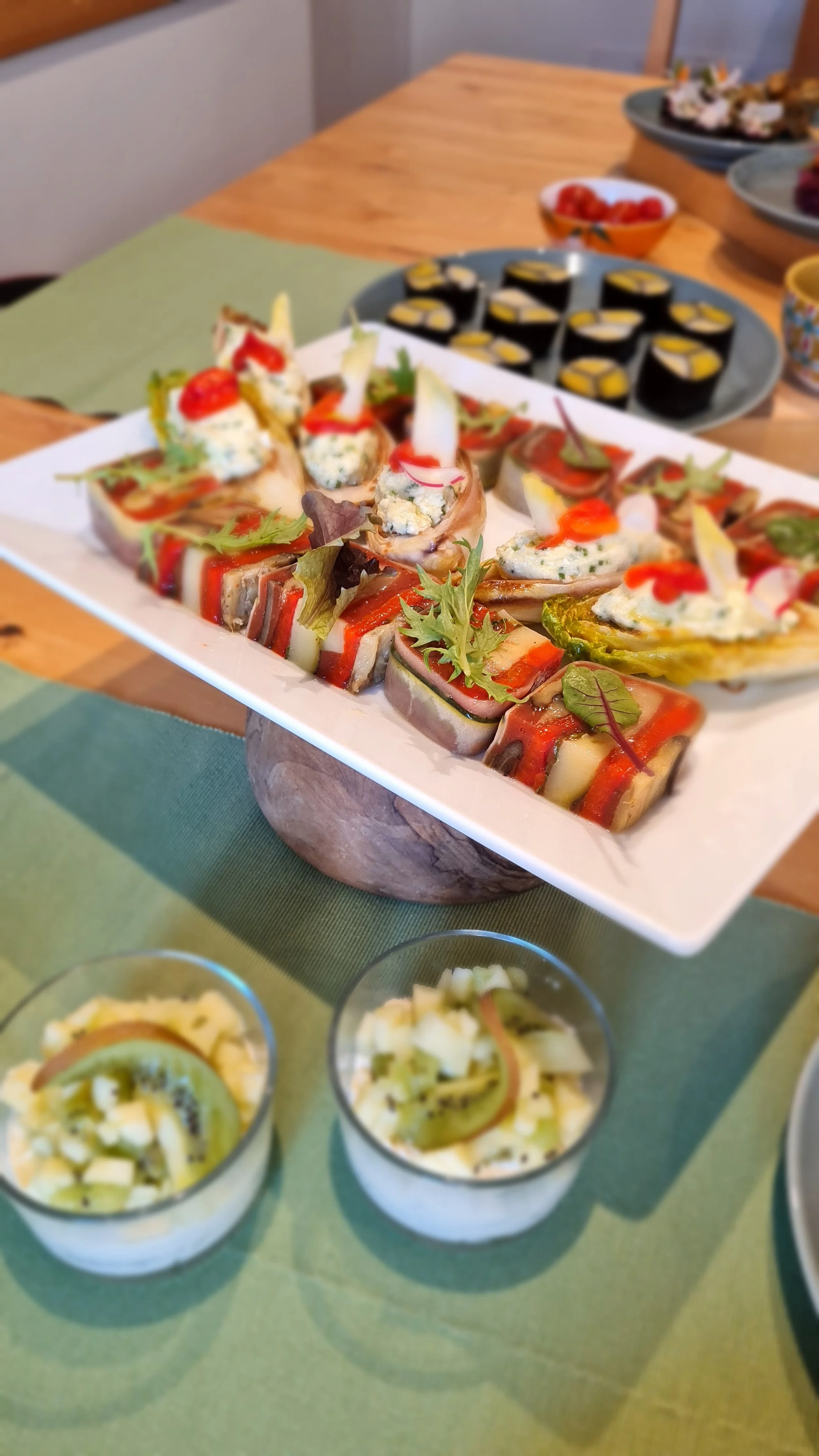 Assorted appetizers including sushi, sushi rolls, and small dessert cups with kiwi and fruit garnishes, arranged on a table.