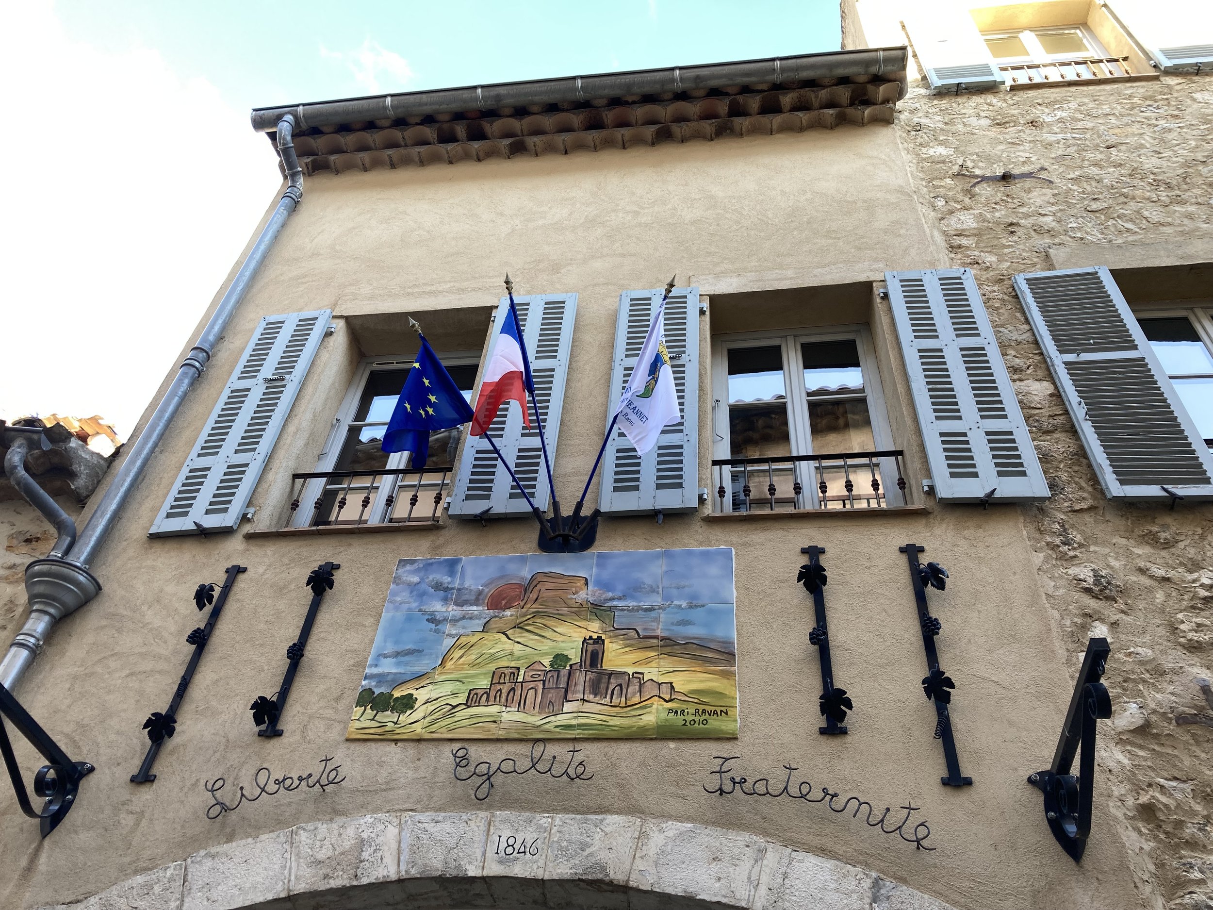 Building facade with three windows with blue shutters, flags of the European Union, France, and a local flag, a colorful tile mural depicting a castle or fortress on a hill, and a script inscription reading "Liberté, Égalité, Fraternité" with the year 1846 below.