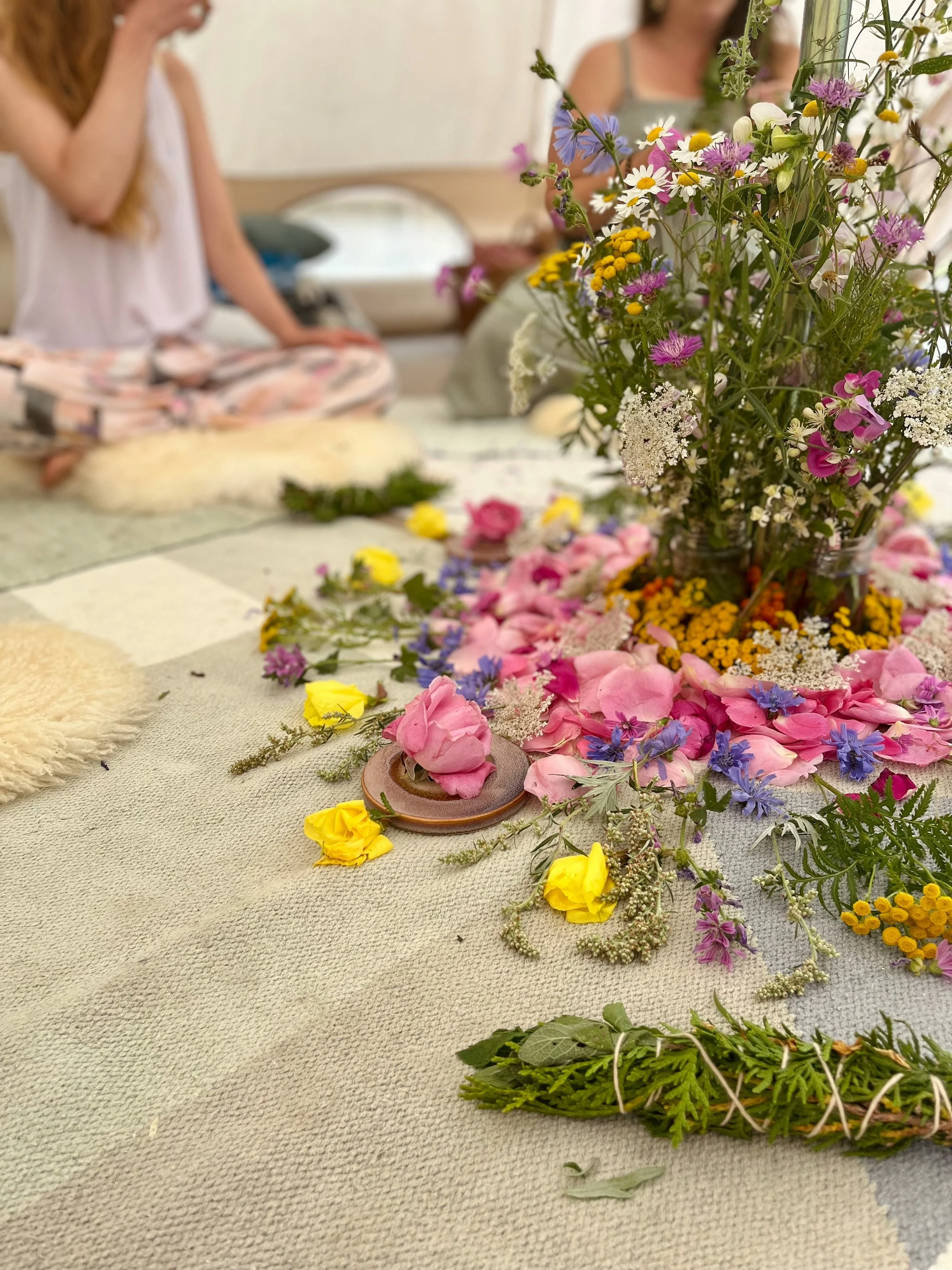 A table decorated with a large bouquet of mixed flowers including pink, purple, yellow, and white blossoms, with petals and greenery scattered on the table. In the background, people are sitting, but their faces are out of focus.