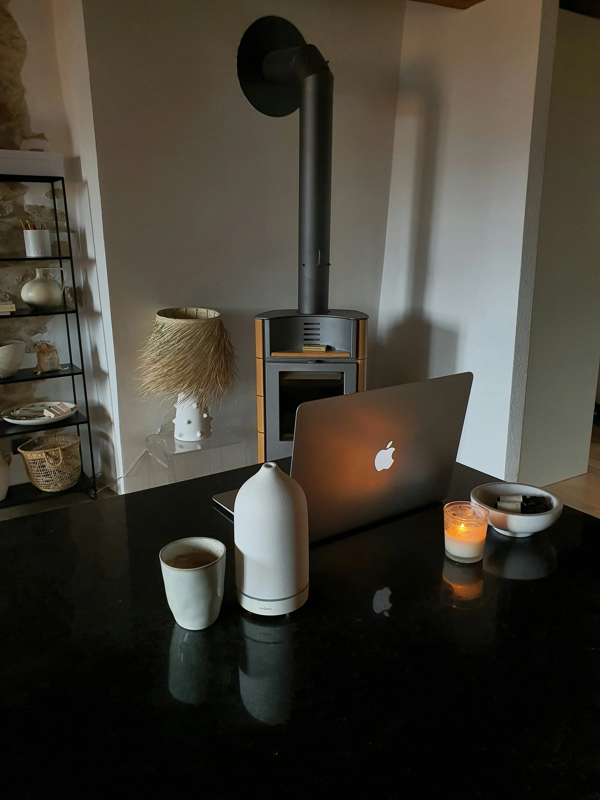 A cozy indoor scene featuring a black countertop with a white cup of coffee, a white electric kettle, a lit candle, and an Apple laptop. In the background, there's a wood stove, a tall black vent pipe, a vintage-style lamp with a feathered shade, and a shelf with decorative items.