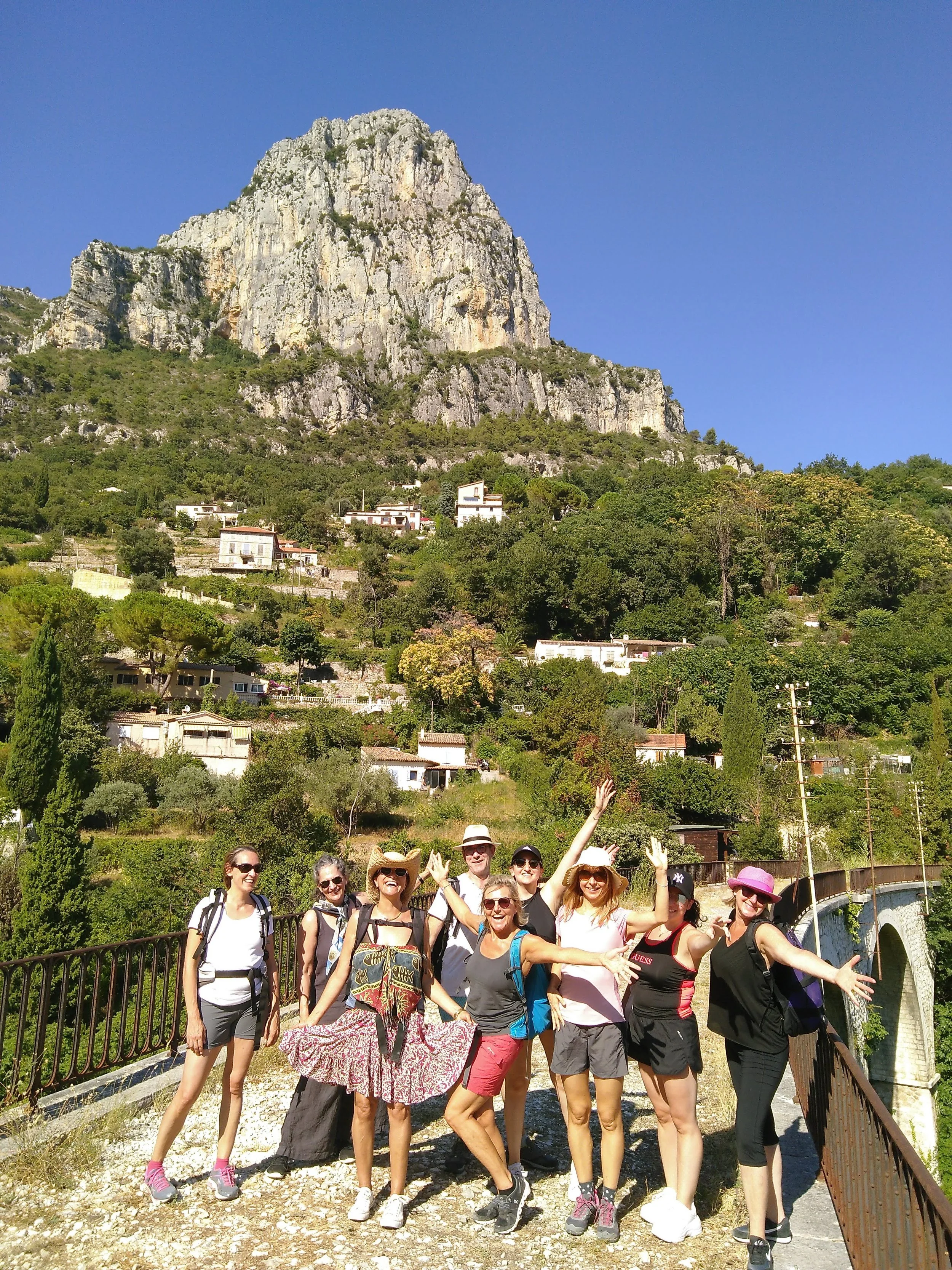 Group of eight women in casual hiking clothes and hats, smiling and posing on a bridge with a scenic mountainous landscape and houses in the background.