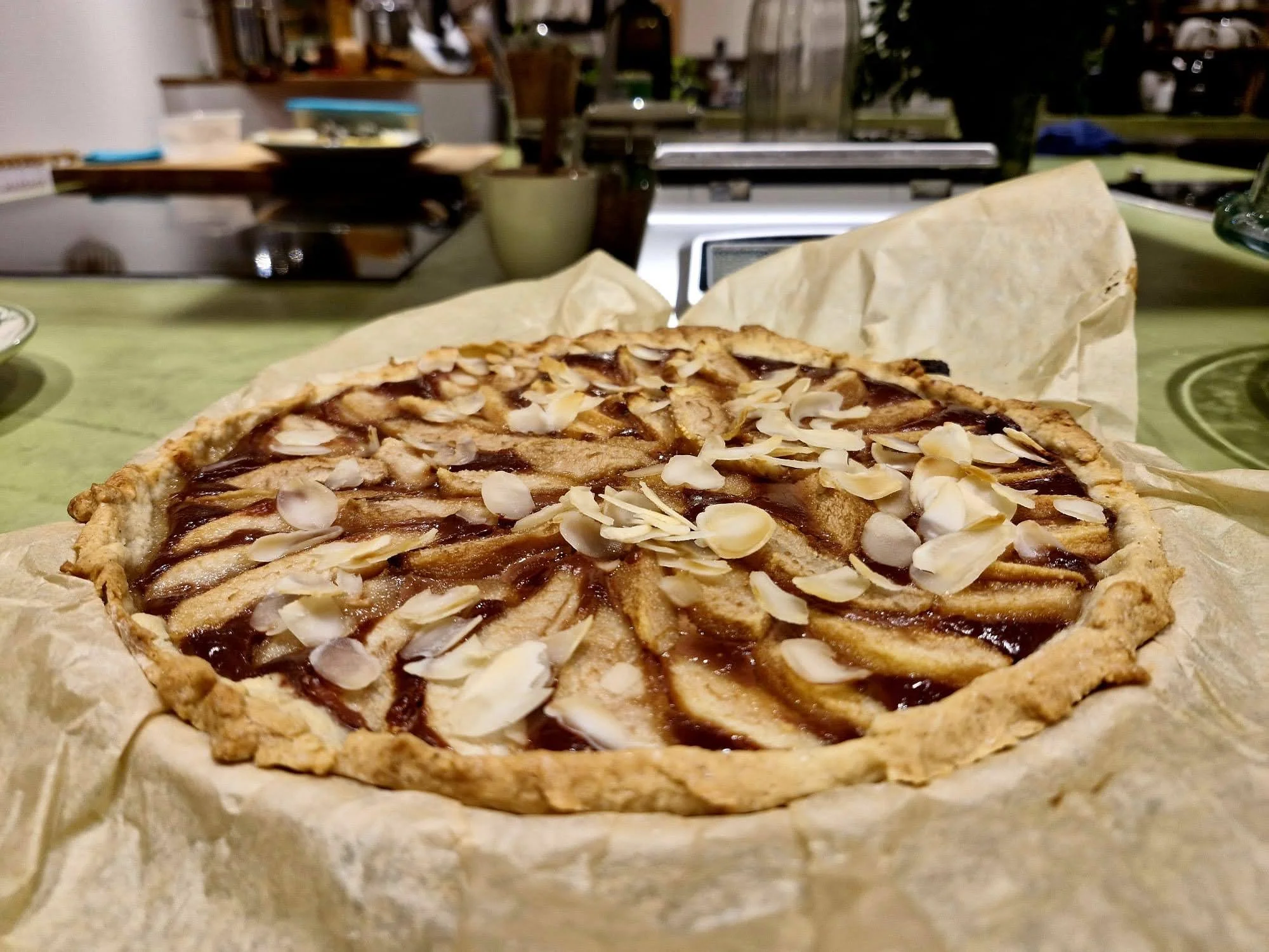A homemade pear tart with sliced pears, almond slices on top, and a glossy glaze, placed on parchment paper on a green kitchen table.