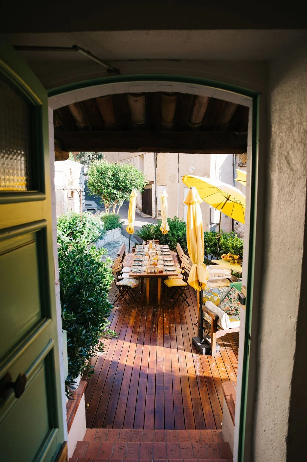View of an outdoor patio with a long dining table set with plates, glasses, and cutlery, surrounded by chairs. Yellow umbrellas provide shade. The patio is paved with wooden planks and greenery, with bushes and a tree in the background.