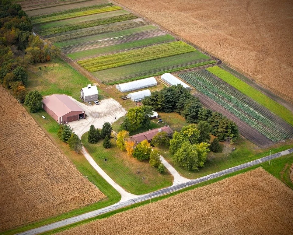 Aerial view of a rural farm with greenhouses, barn, house, and cultivated fields surrounded by farmland.