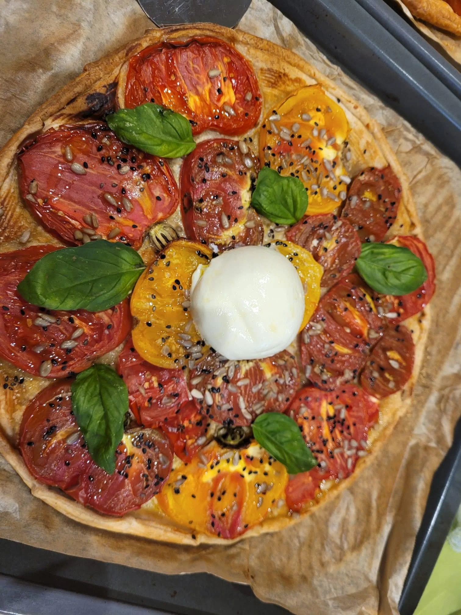 A tomato tart with yellow and red heirloom tomatoes, fresh basil leaves, and a dollop of burrata cheese, sprinkled with black sesame seeds and sunflower seeds.