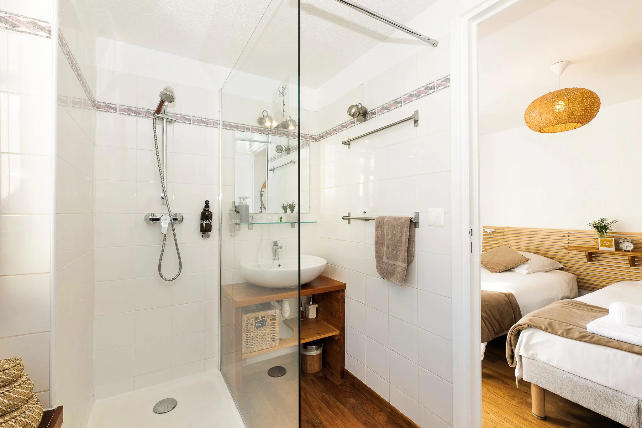 Bathroom with a walk-in shower, white tiled walls, and a mirror, with view into a bedroom with two beds and wooden accents.