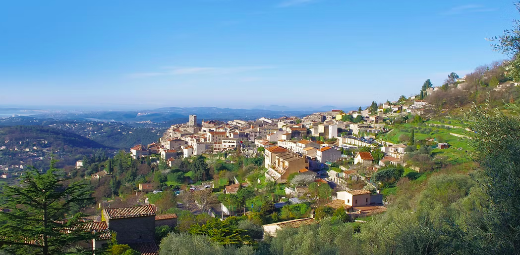 A hillside town with tightly packed buildings overlooking a distant view of the bay and horizon under a clear blue sky.