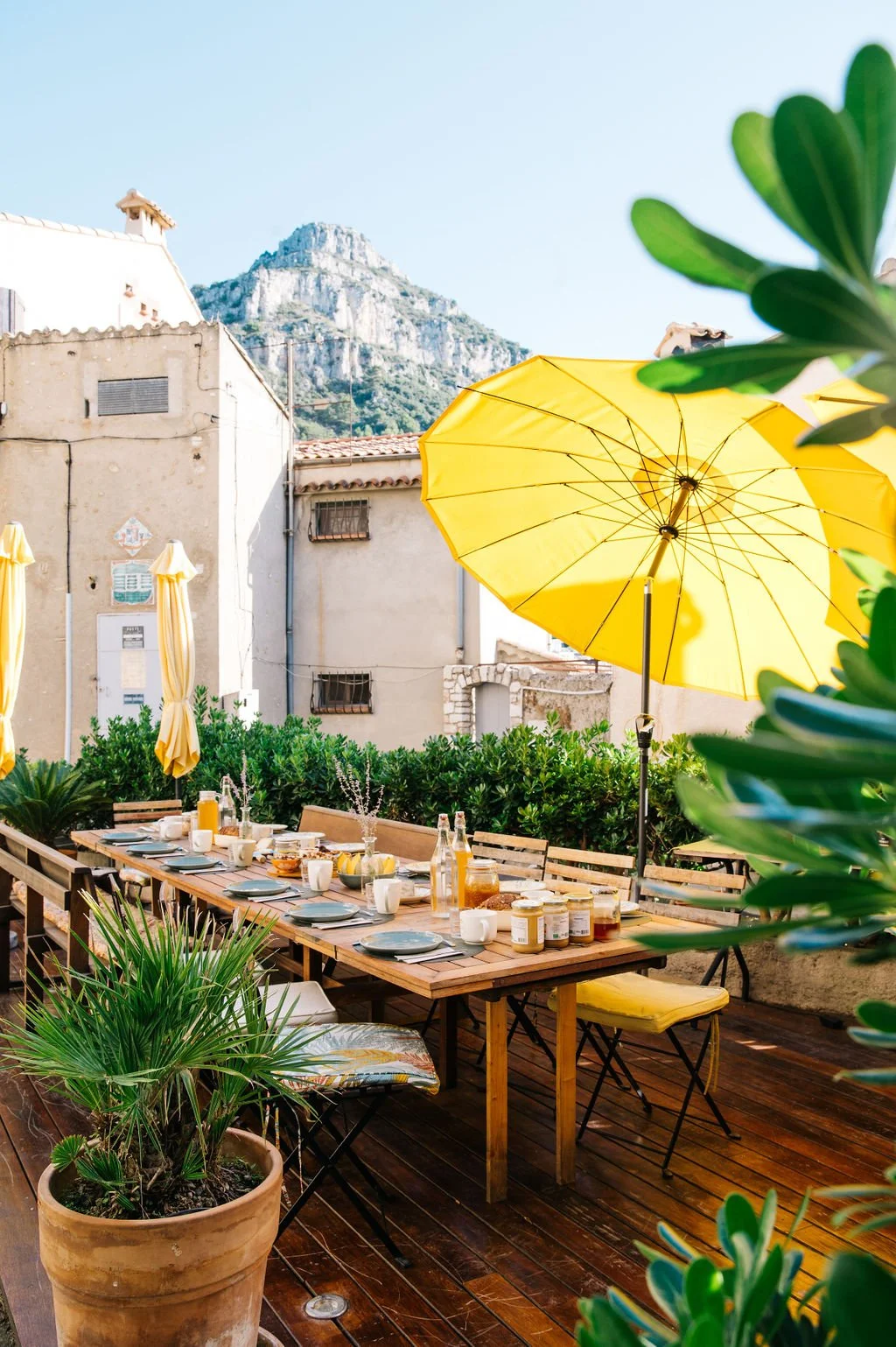 An outdoor dining area on a wooden deck with a long table set for breakfast, featuring plates, cups, jars, and a glass of orange juice. A large yellow umbrella provides shade, and there is green foliage and potted plants around. In the background, there are old buildings and a mountain under a clear blue sky.