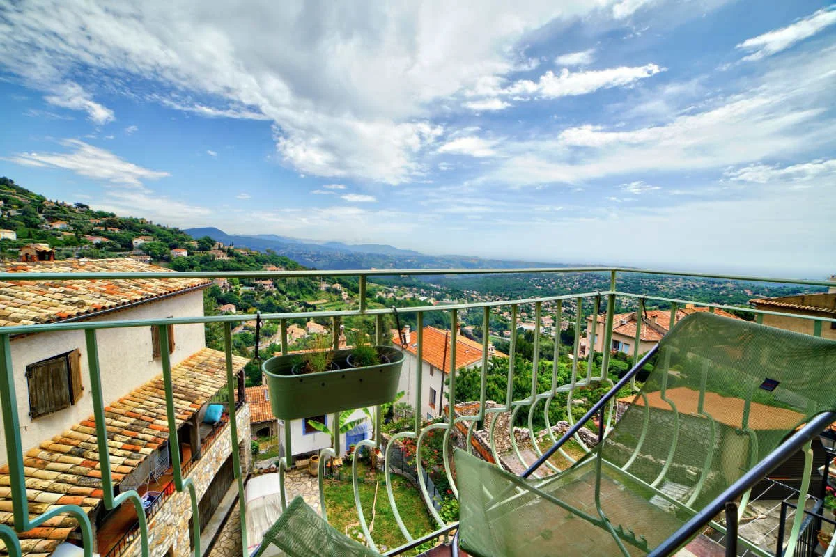 View from a balcony overlooking a hilly landscape with houses, greenery, and distant mountains under a partly cloudy sky.