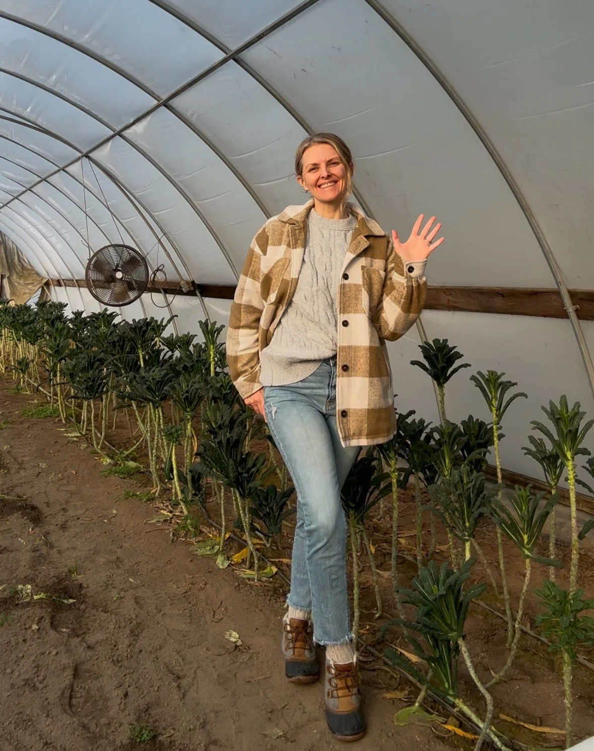 Woman smiling and waving inside a greenhouse, standing next to planted vegetables or herbs, wearing a plaid jacket and jeans.