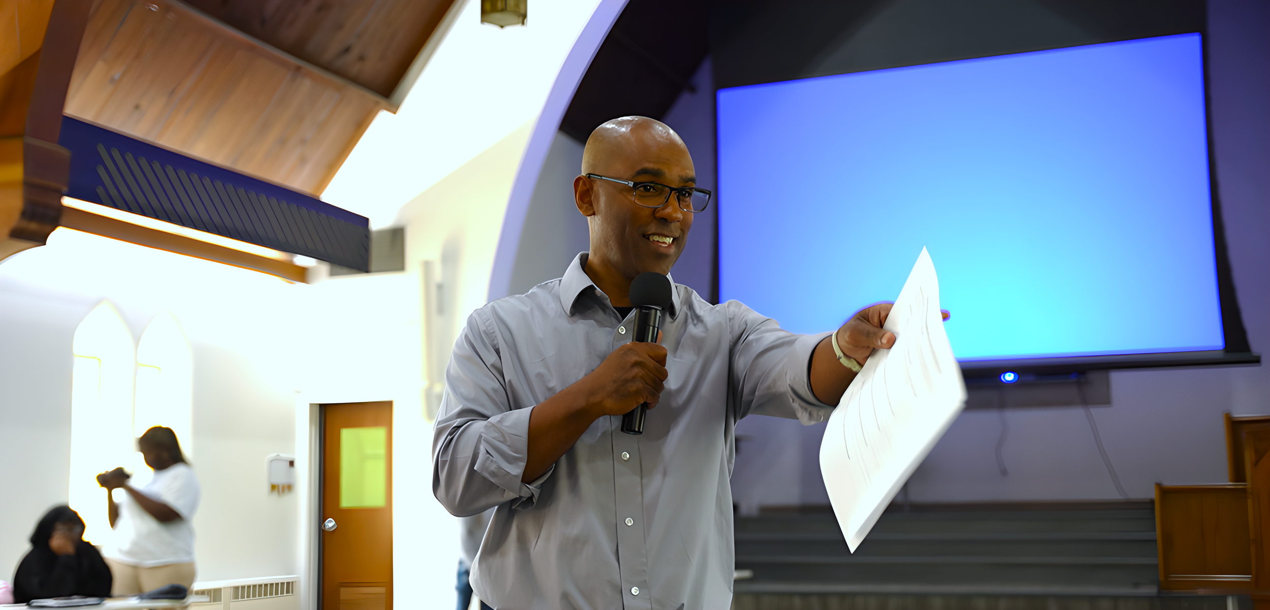A man with glasses, wearing a light gray shirt, holding a microphone in one hand and a sheet of paper in the other, speaking in front of a large blank screen in a room with white walls and wooden accents.