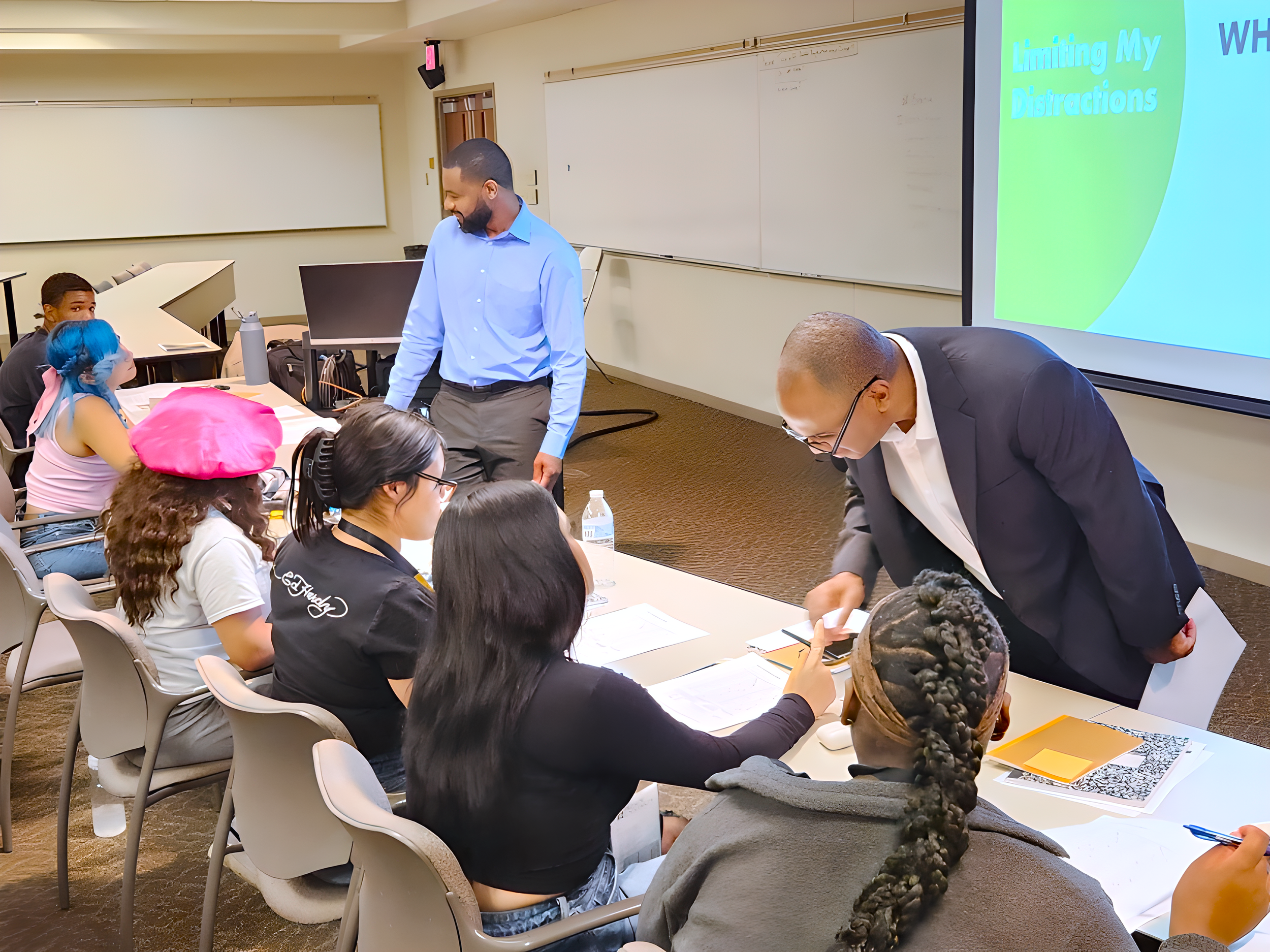 A group of students seated at a table in a classroom or conference room, with two men standing and interacting with the students. There is a presentation slide on the screen that reads 'Limiting My Distractions'.