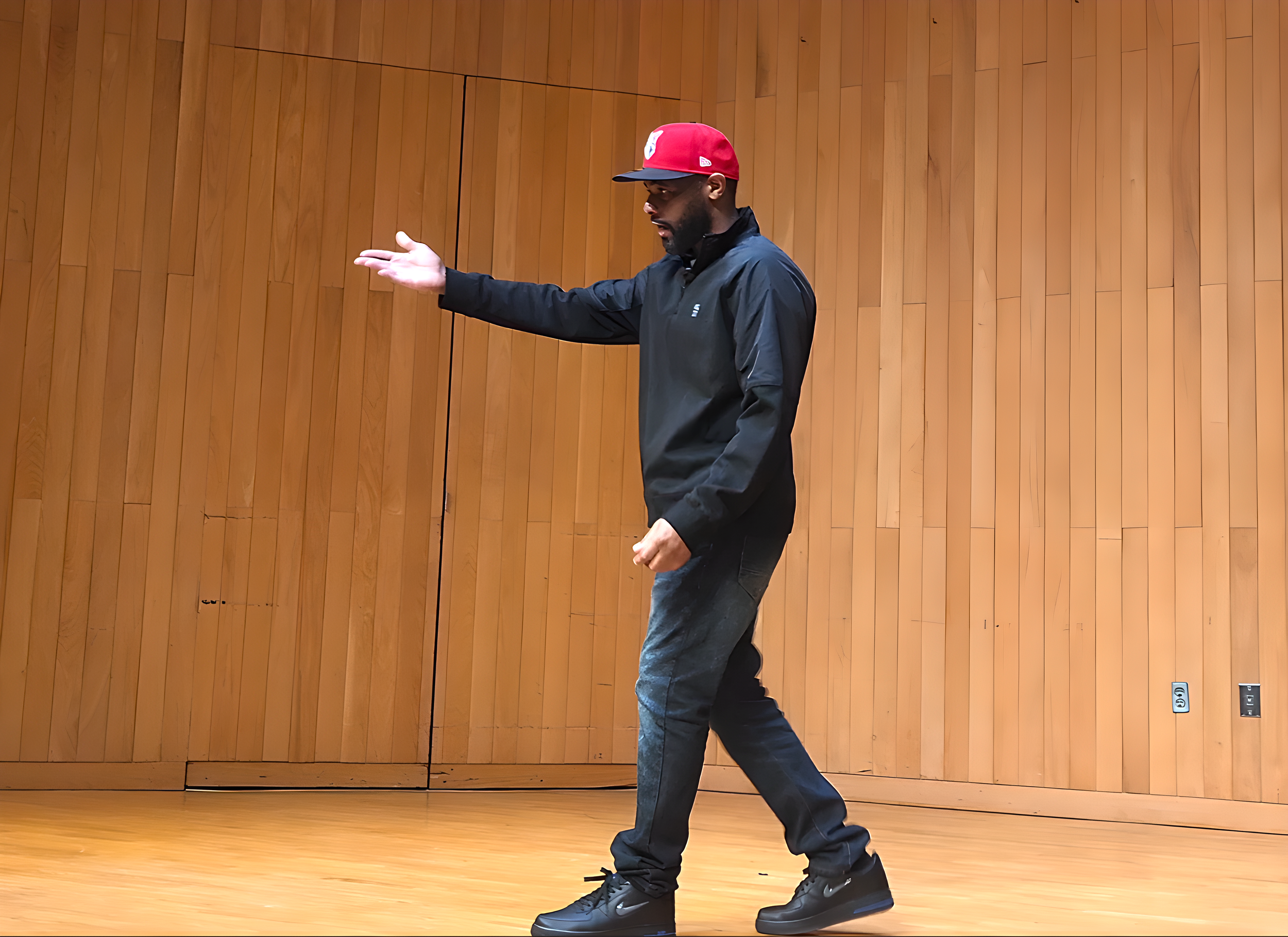 A man wearing a red baseball cap, black jacket, jeans, and black shoes, standing on a wooden floor in front of a wooden-paneled wall, with his right arm extended outward.