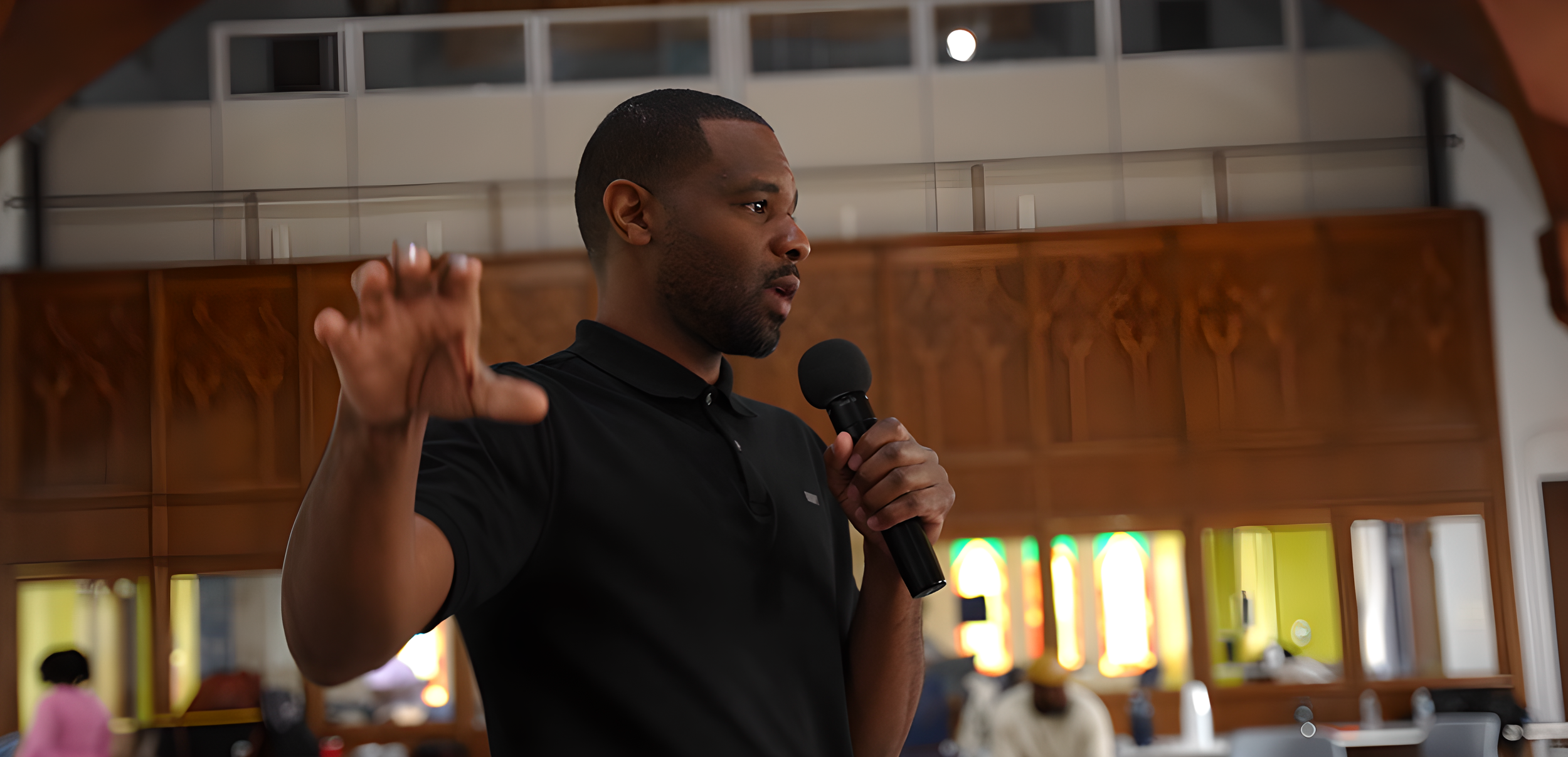 Man in black polo shirt speaking into a microphone inside a room with wooden decor and stained glass windows.