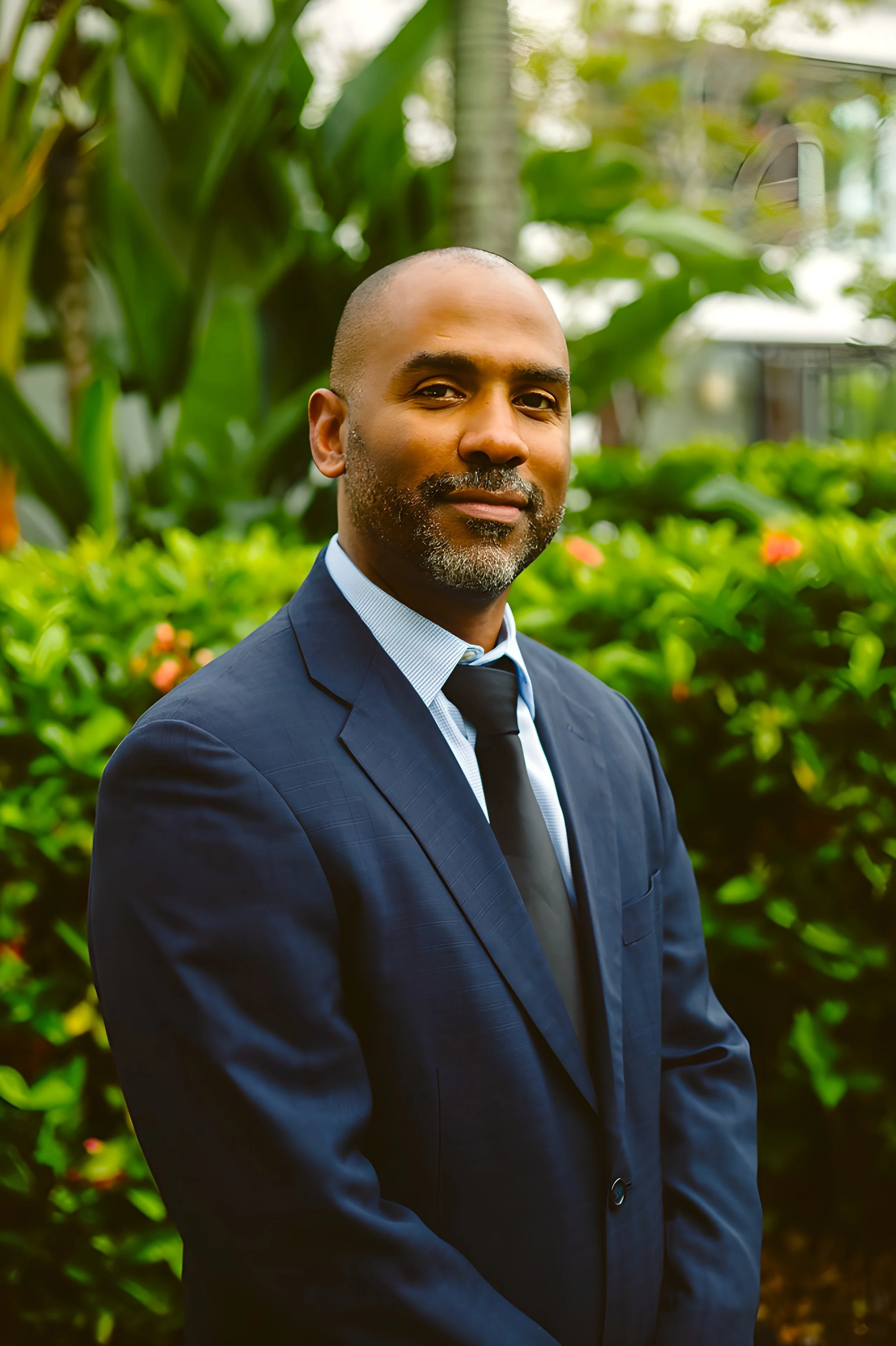 Portrait of a man wearing a dark blue suit, light blue shirt, and black tie, standing outdoors with green foliage and plants in the background.