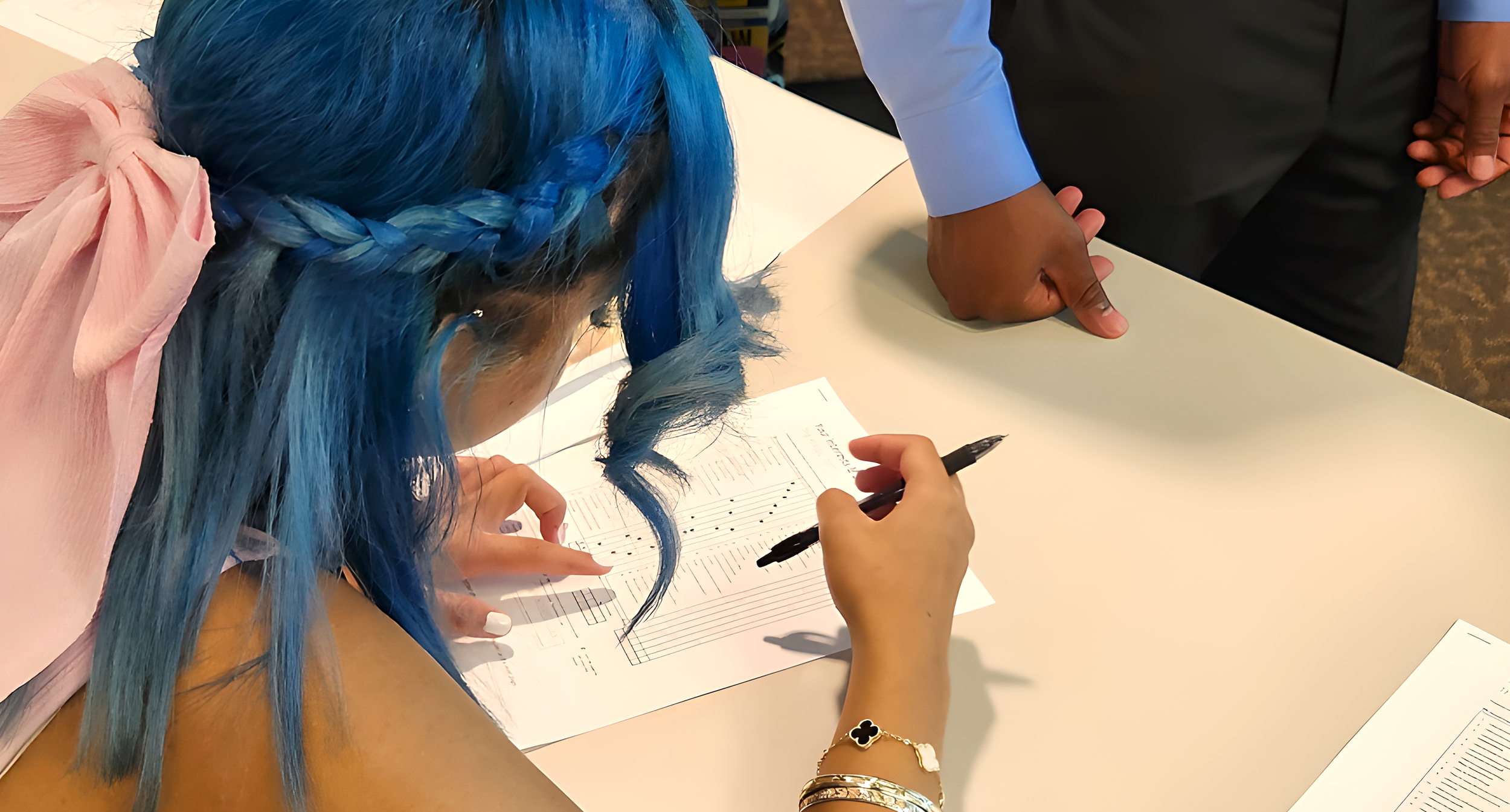 Person with blue hair signing sheet music on a table while another person stands nearby
