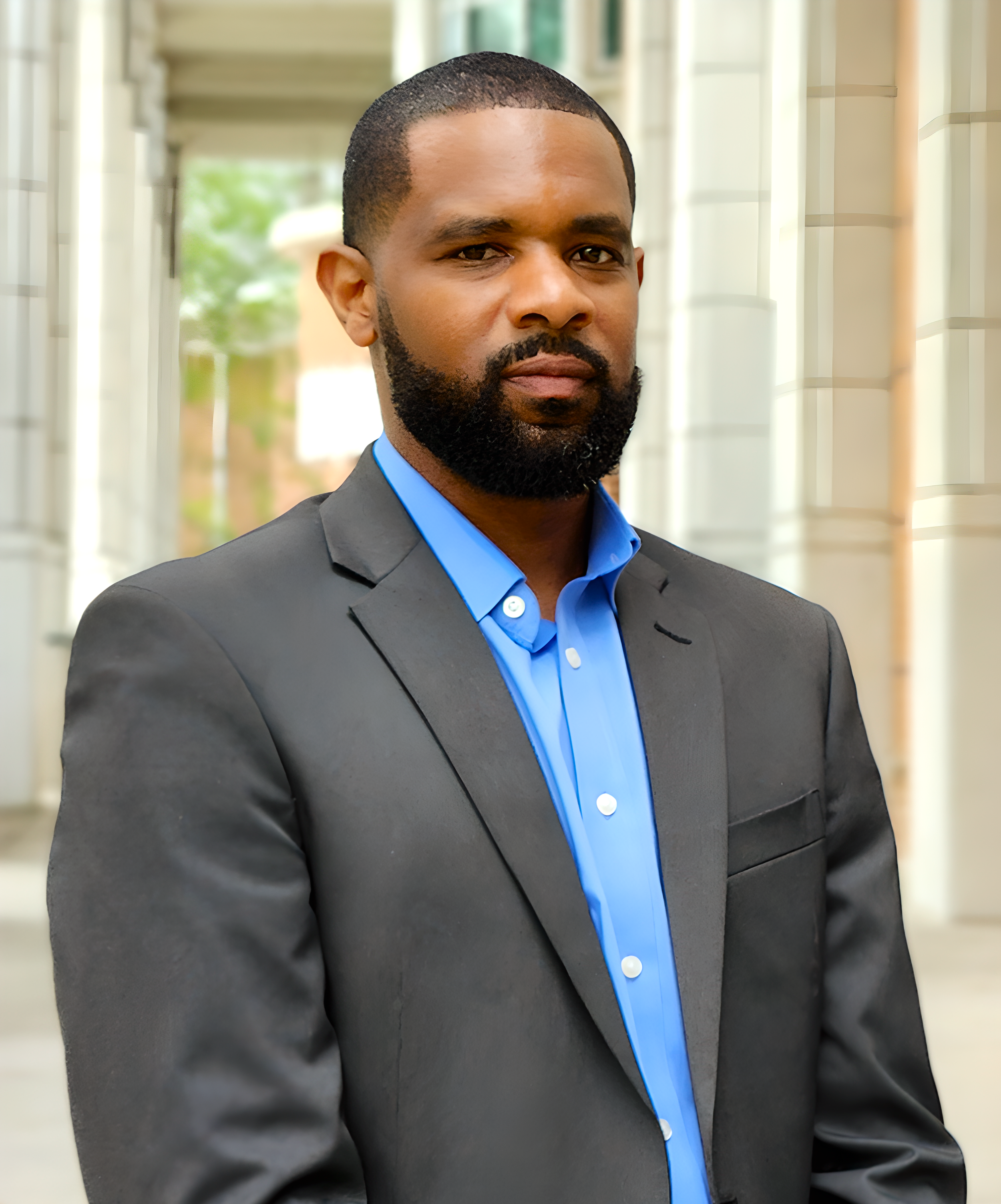 A man in a gray suit and blue shirt standing outdoors in front of a modern building.