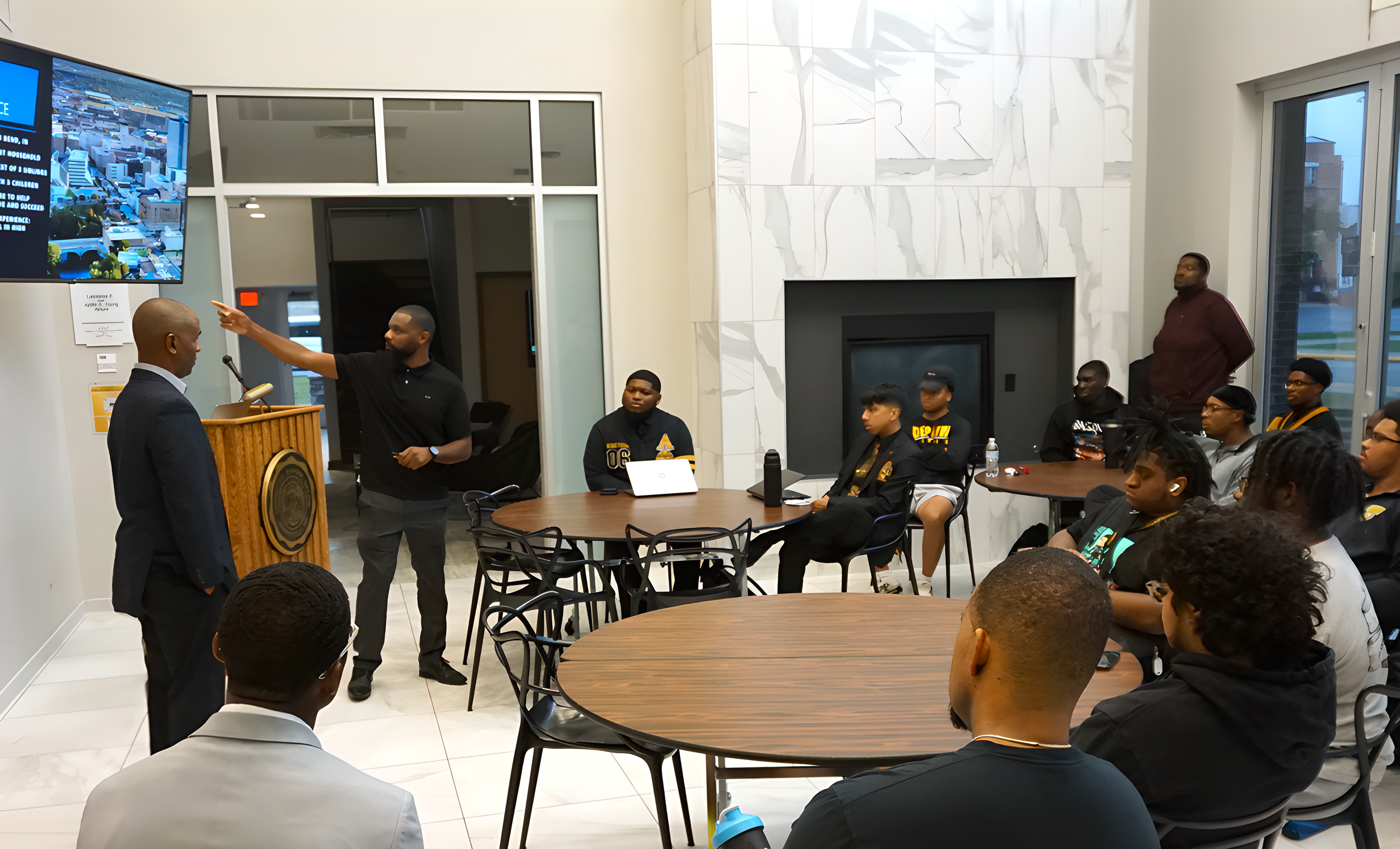 A man in a suit speaking at a podium while another man points at a large screen displaying an aerial view of a city. Several people are seated around tables, listening attentively in a modern room with white marble walls and large windows.