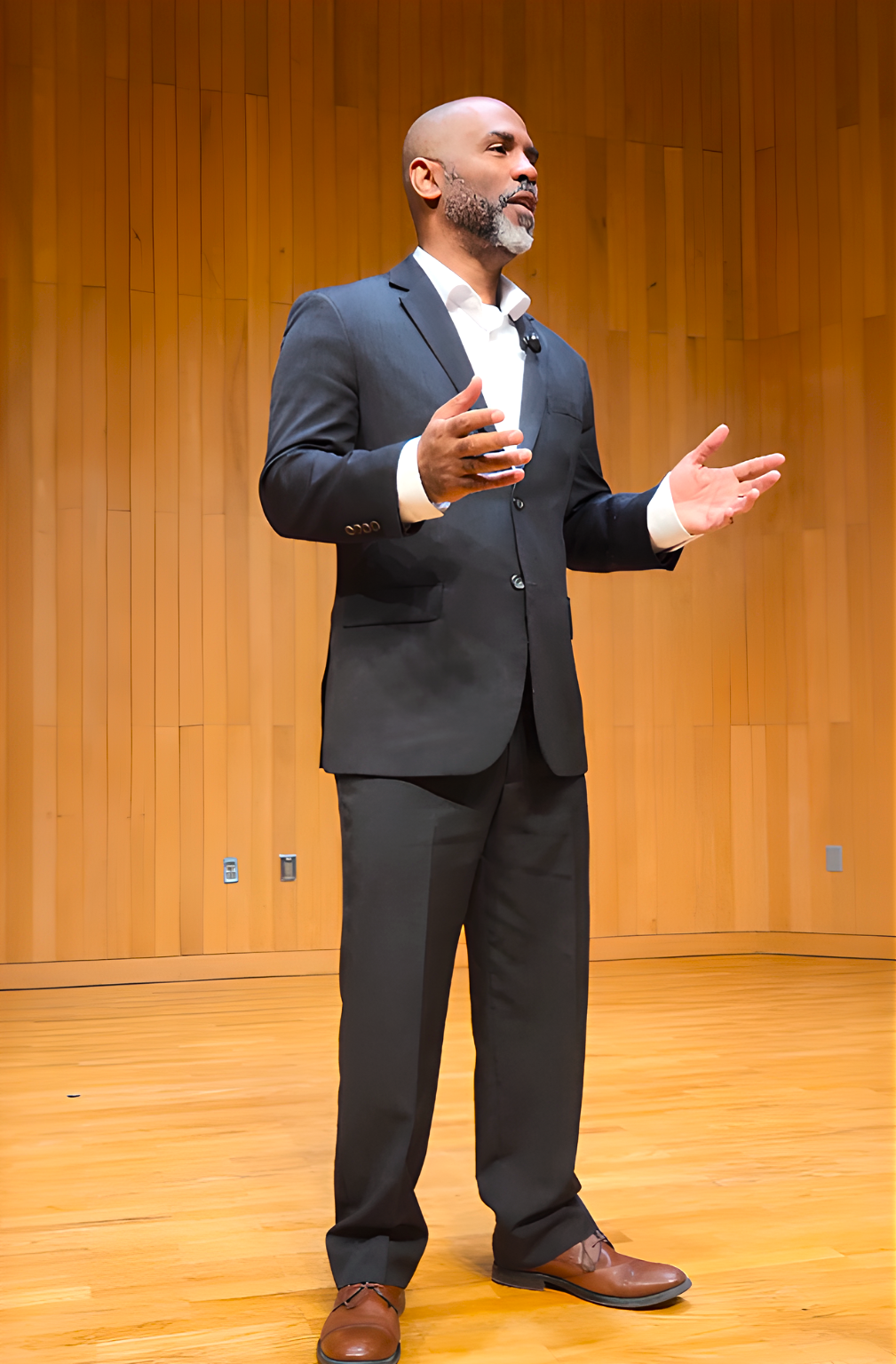 A man in a dark suit speaking on stage in front of a wooden wall.