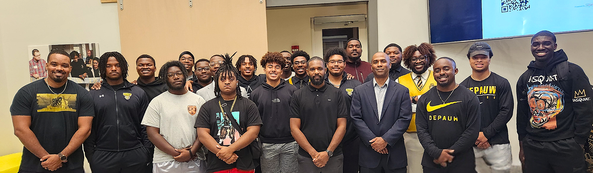 A group of young people and adults posing for a photo in an indoor setting. Some are wearing casual clothing, others in sportswear or suits. They are smiling and standing close together.