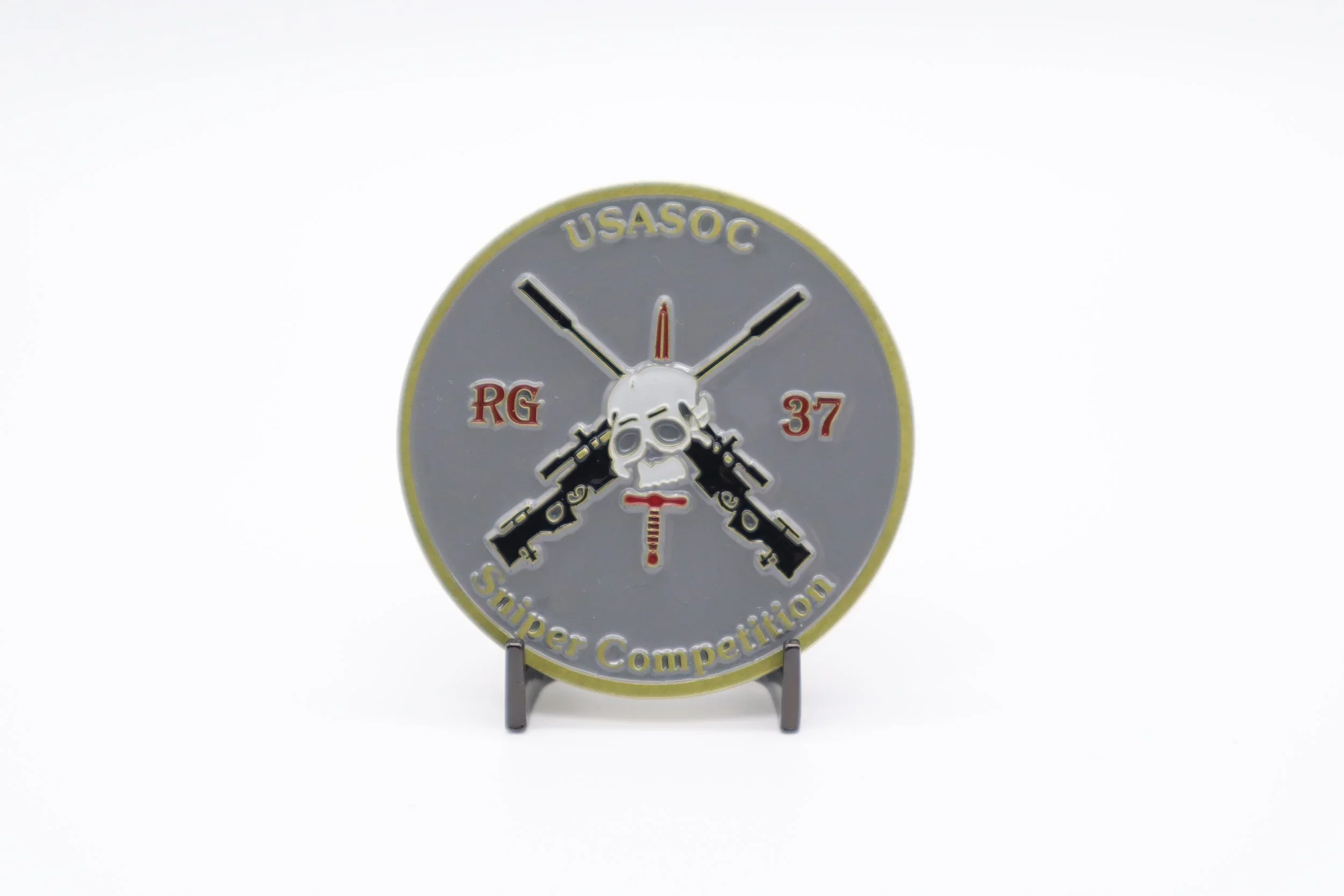 A circular badge with a skull and crossed rifles in the center, with the text 'USASOC', 'RG 37', and 'Sniper Competition' around the edge, displayed on a stand.