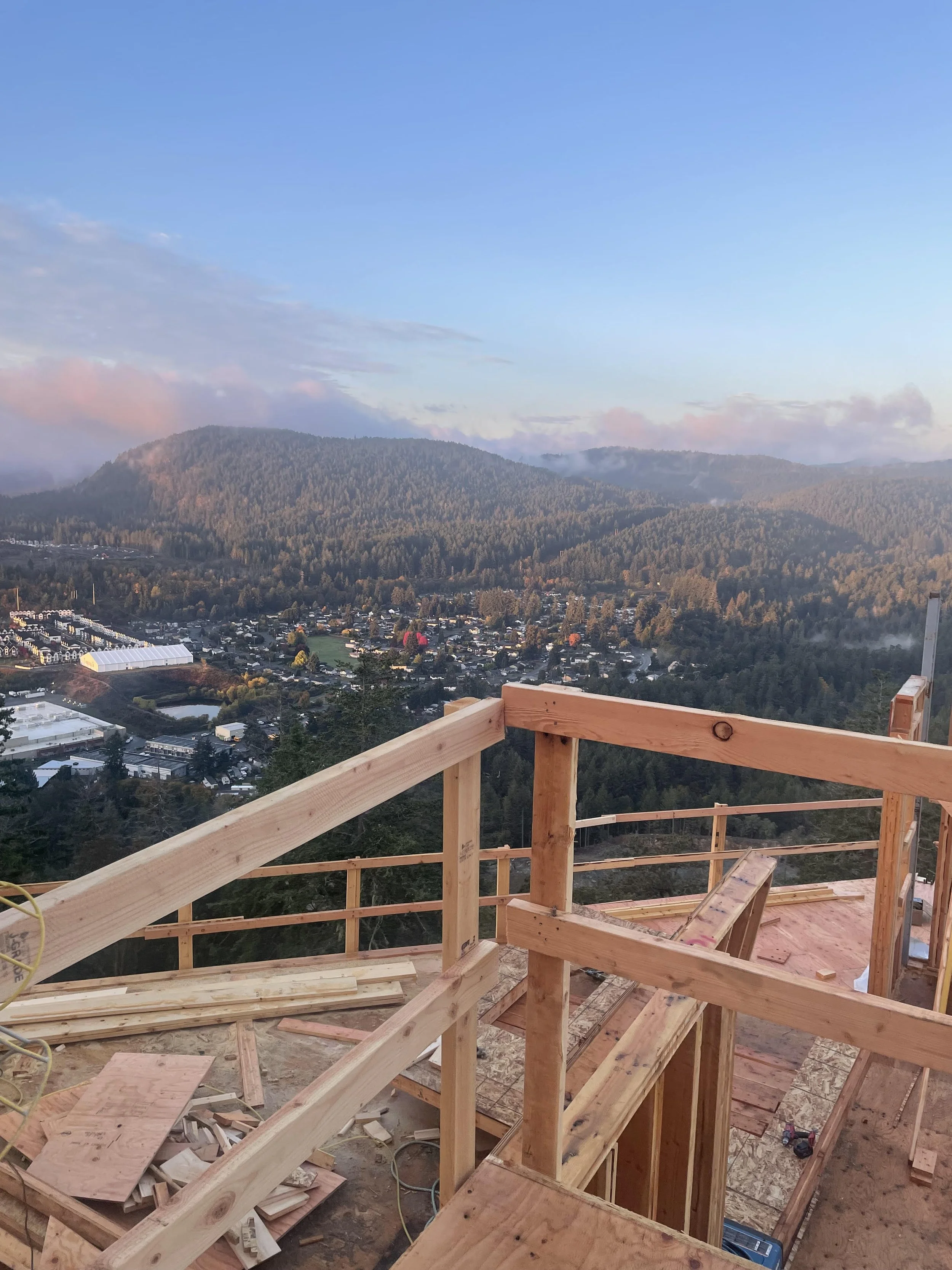 View of a construction site with a wooden frame overlooking a town and mountains in the distance under a blue sky.