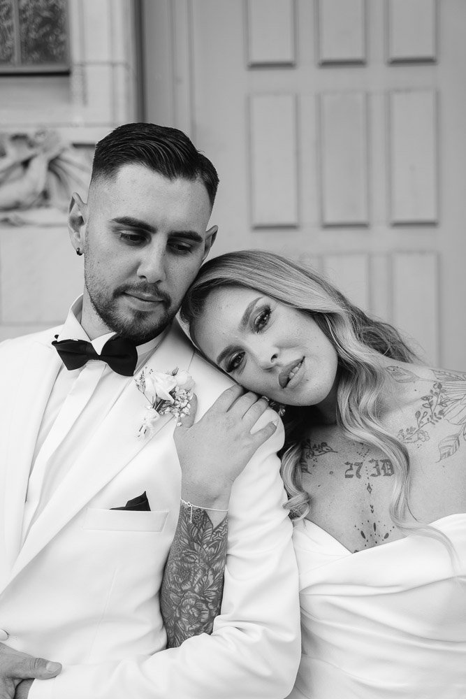Ein junges Paar an einem Hochzeitstag, der Mann trägt einen weißen Anzug mit Fliege, die Frau trägt ein weißes Kleid mit Tattoos auf Schultern und Armen, sie sitzen eng beieinander, die Frau lehnt ihren Kopf an die Schulter des Mannes.
