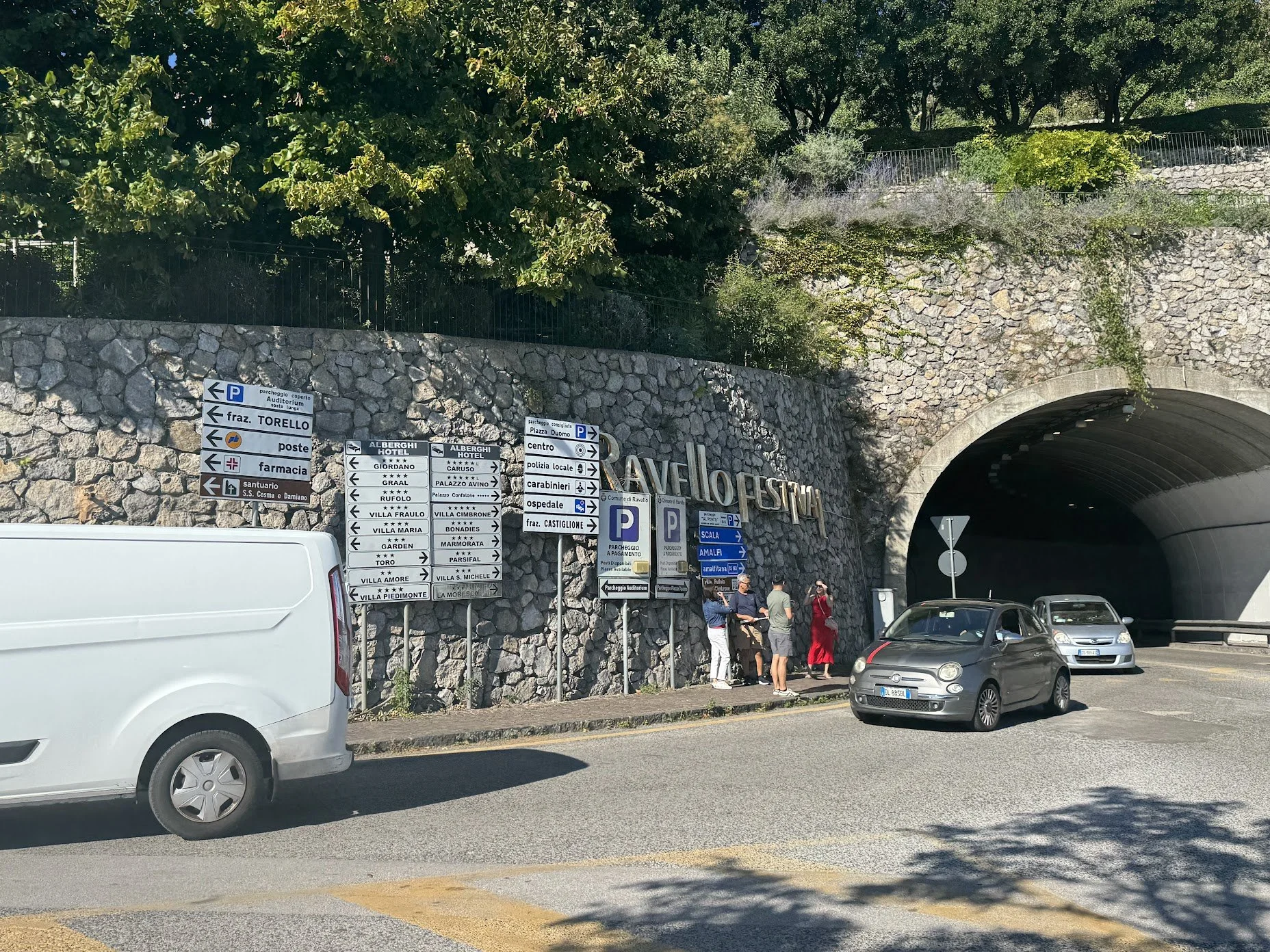 People waiting at a tunnel entrance with numerous directed signs, parked cars, greenery on the hillside above, and shadows on the road.