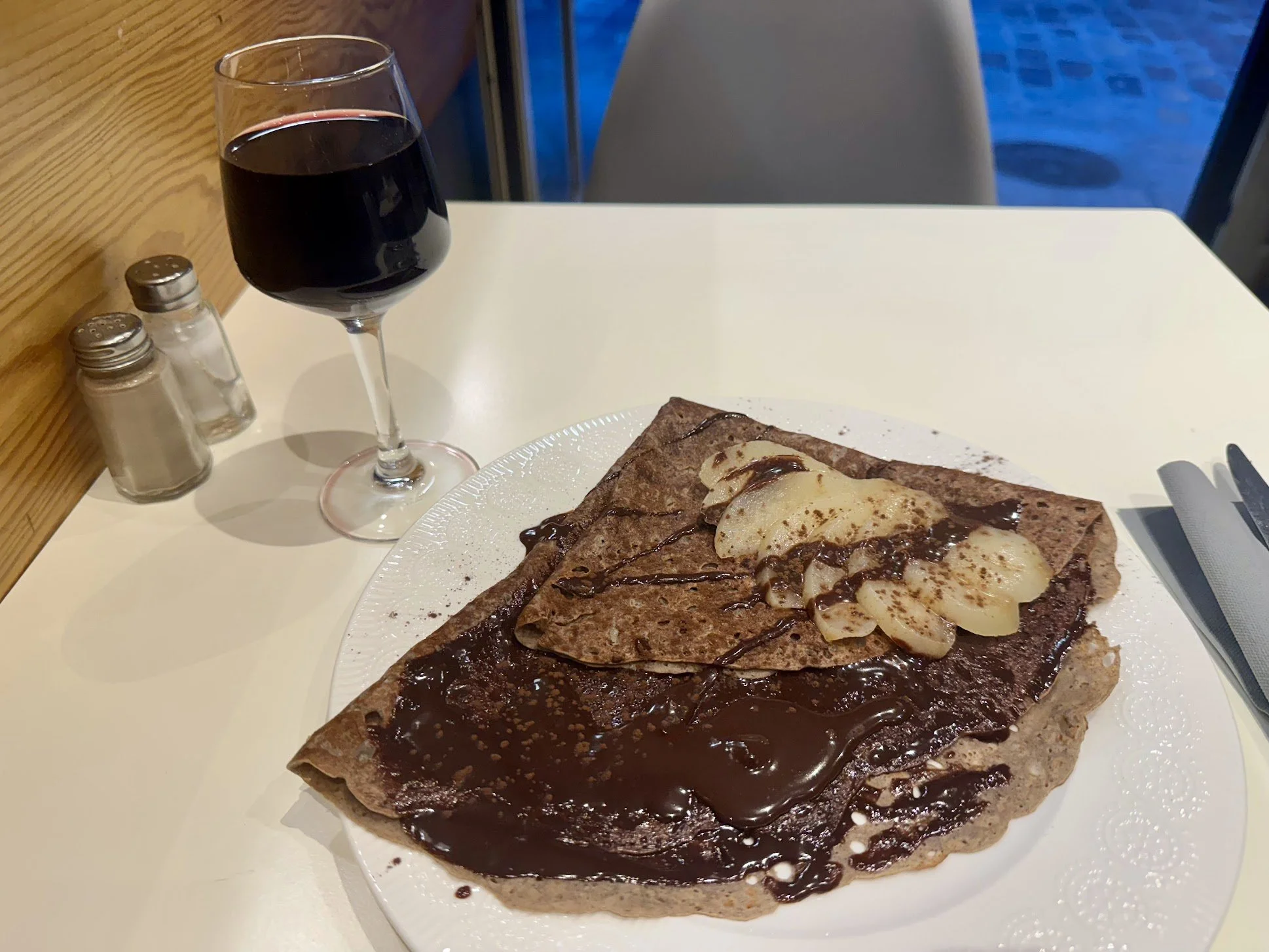 Plate of crepe with bananas and chocolate, glass of red wine, salt and pepper shakers on a white table.