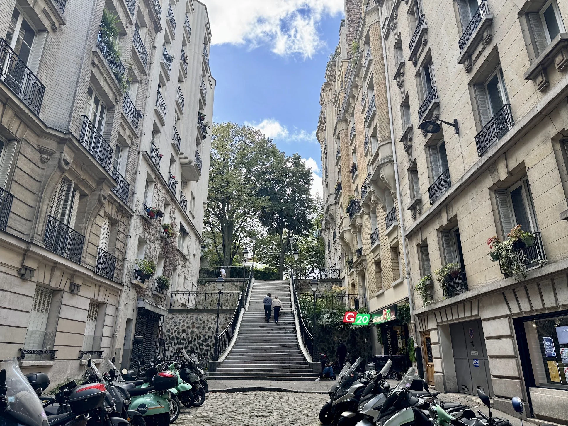 City street scene in Paris with stairs between tall buildings, parked scooters in the foreground, and a tree at the top of the stairs under a partly cloudy sky.
