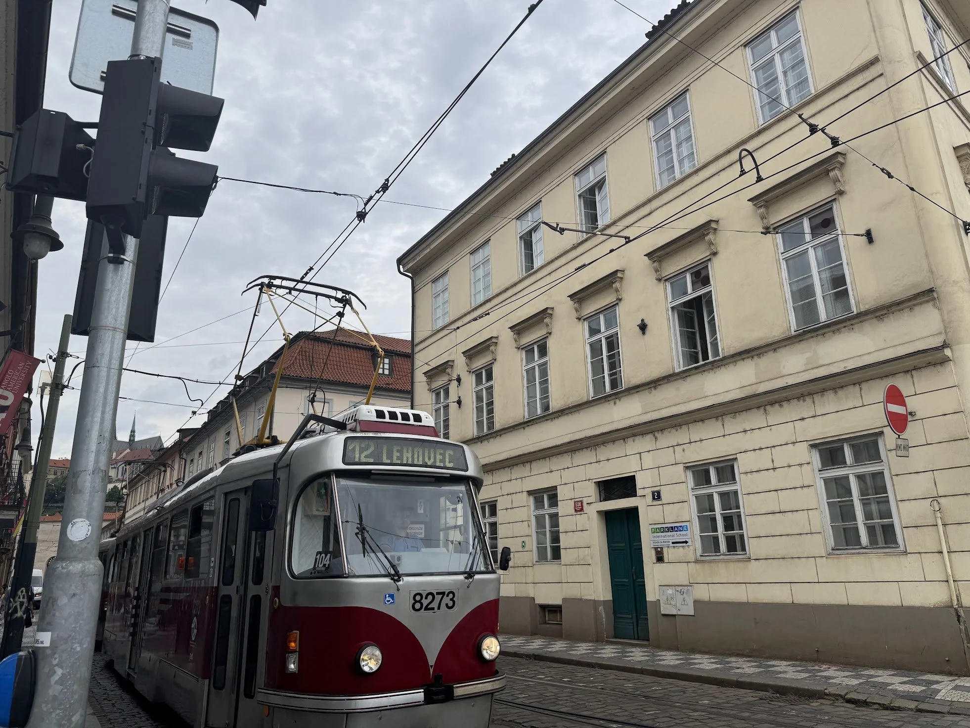 City street with tramcar, overhead wires, traffic light, residential buildings, and cloudy sky.
