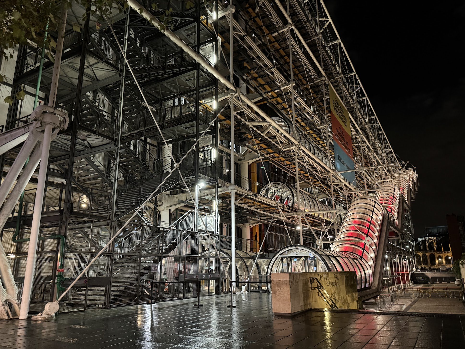 Night view of a modern urban building with exposed metal framework, staircases, and a glass tube structure illuminated by lights, with a sign hanging from the upper part.
