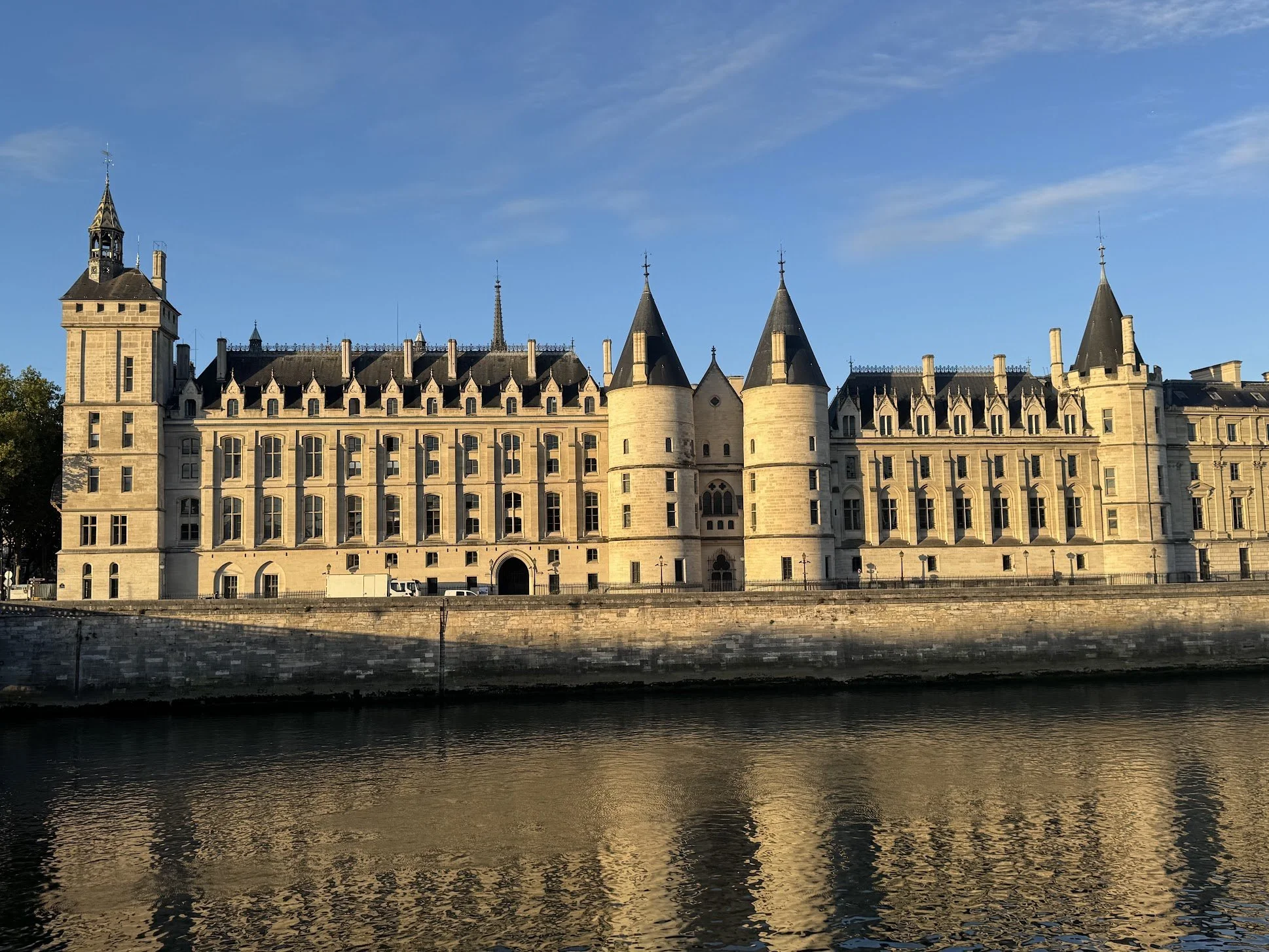 A historic castle with multiple towers and spires, built with light-colored stone, situated along a river with a stone embankment, under a clear blue sky.