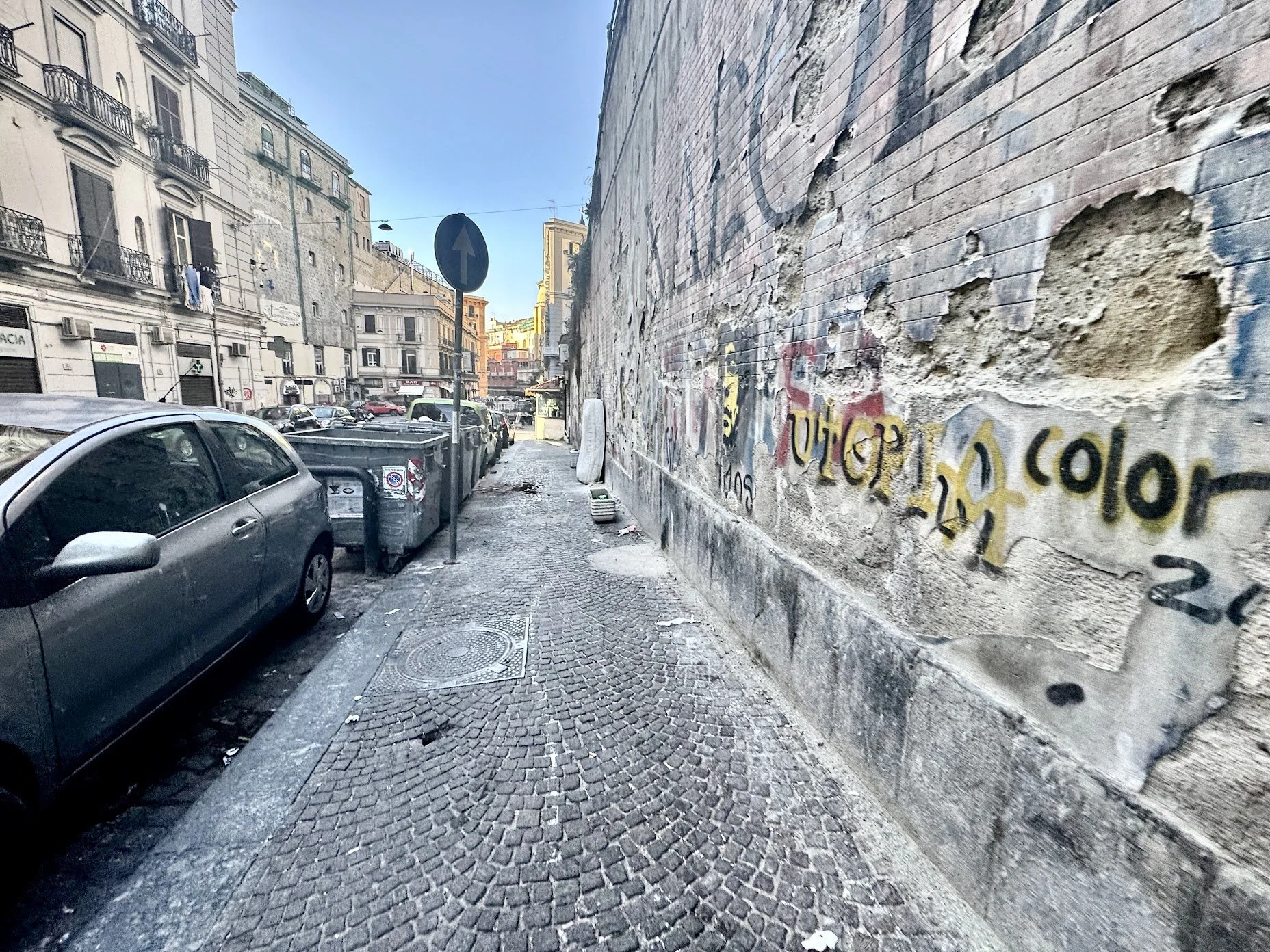 City street scene with parked cars along a sidewalk, graffiti-covered weathered brick wall, buildings with balconies, and a traffic sign against a blue sky.