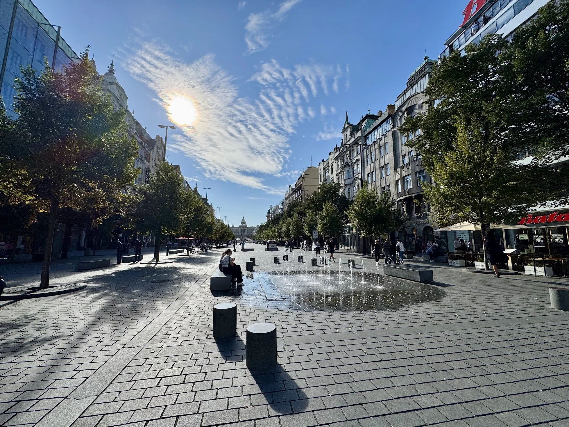 An urban street scene with a wide pedestrian area, trees, and fountains. Modern buildings line both sides, with a notable dome and clock tower in the distance. The sky is partly cloudy with the sun shining through.