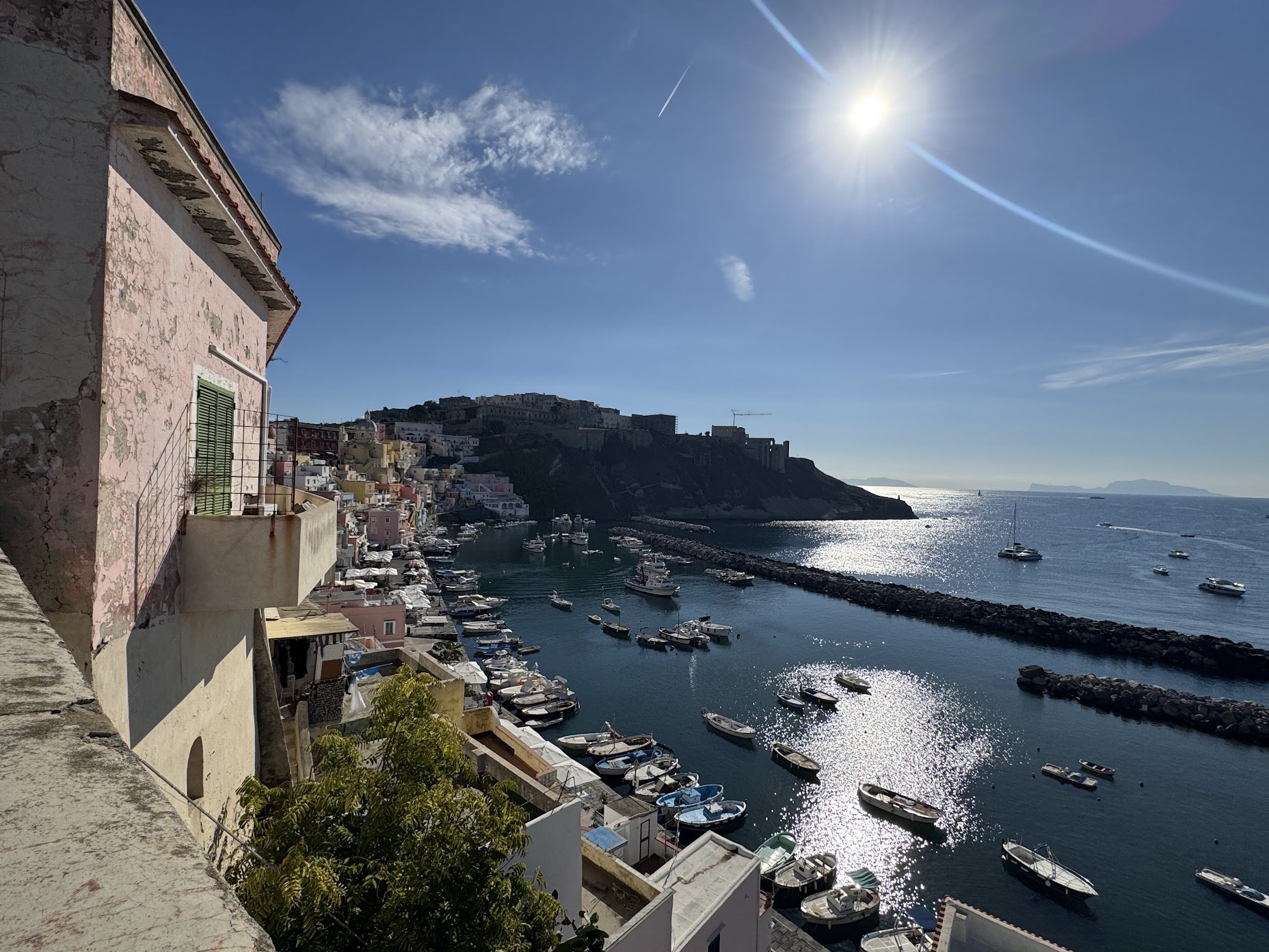 A coastal cityscape featuring a hillside with colorful buildings overlooking a marina filled with boats and yachts under a bright sun and blue sky.