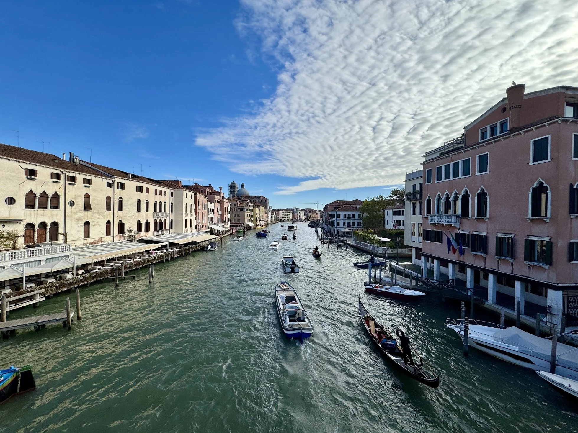 A scenic view of a canal in Venice, Italy, with boats, historic buildings along the water, and a partly cloudy sky.