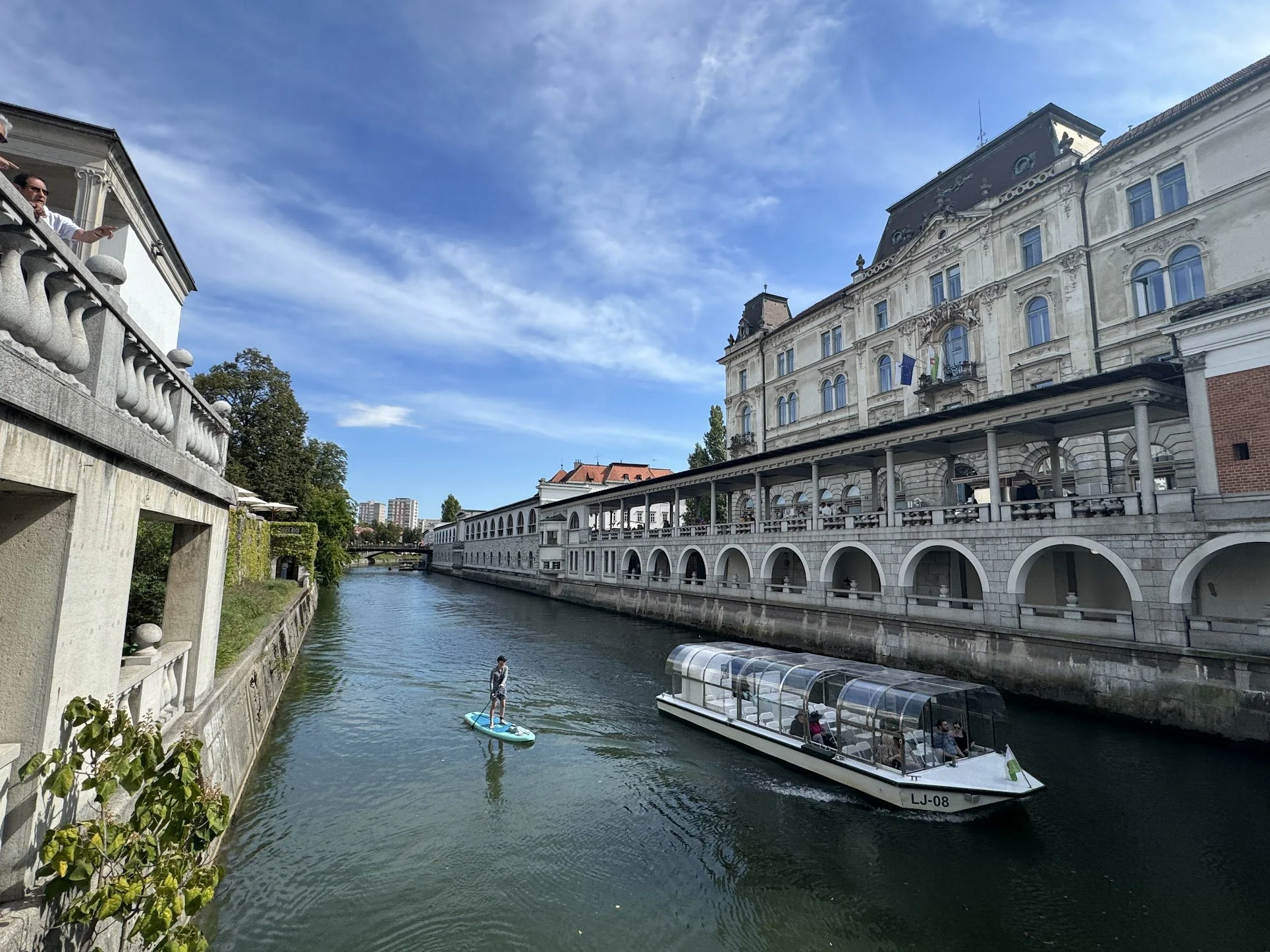 A person paddleboarding on a canal beside historic buildings, with a sightseeing boat carrying passengers passing by in a European city.