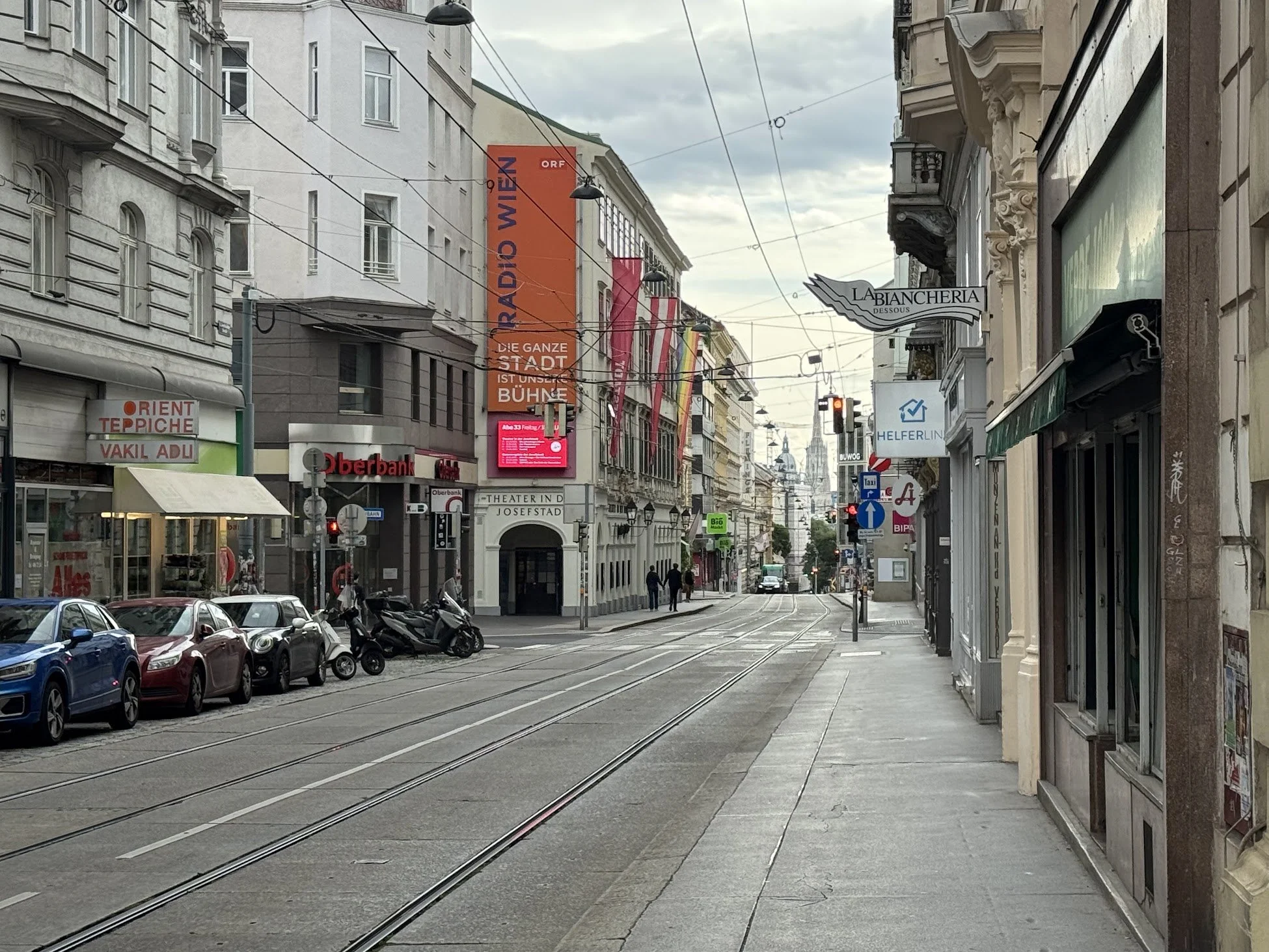 A city street with narrow tracks for tram or trolley cars, cars parked along the curb, and buildings lining both sides. The sky is overcast, and there are signs in German and various businesses visible, including a theater and a bank.