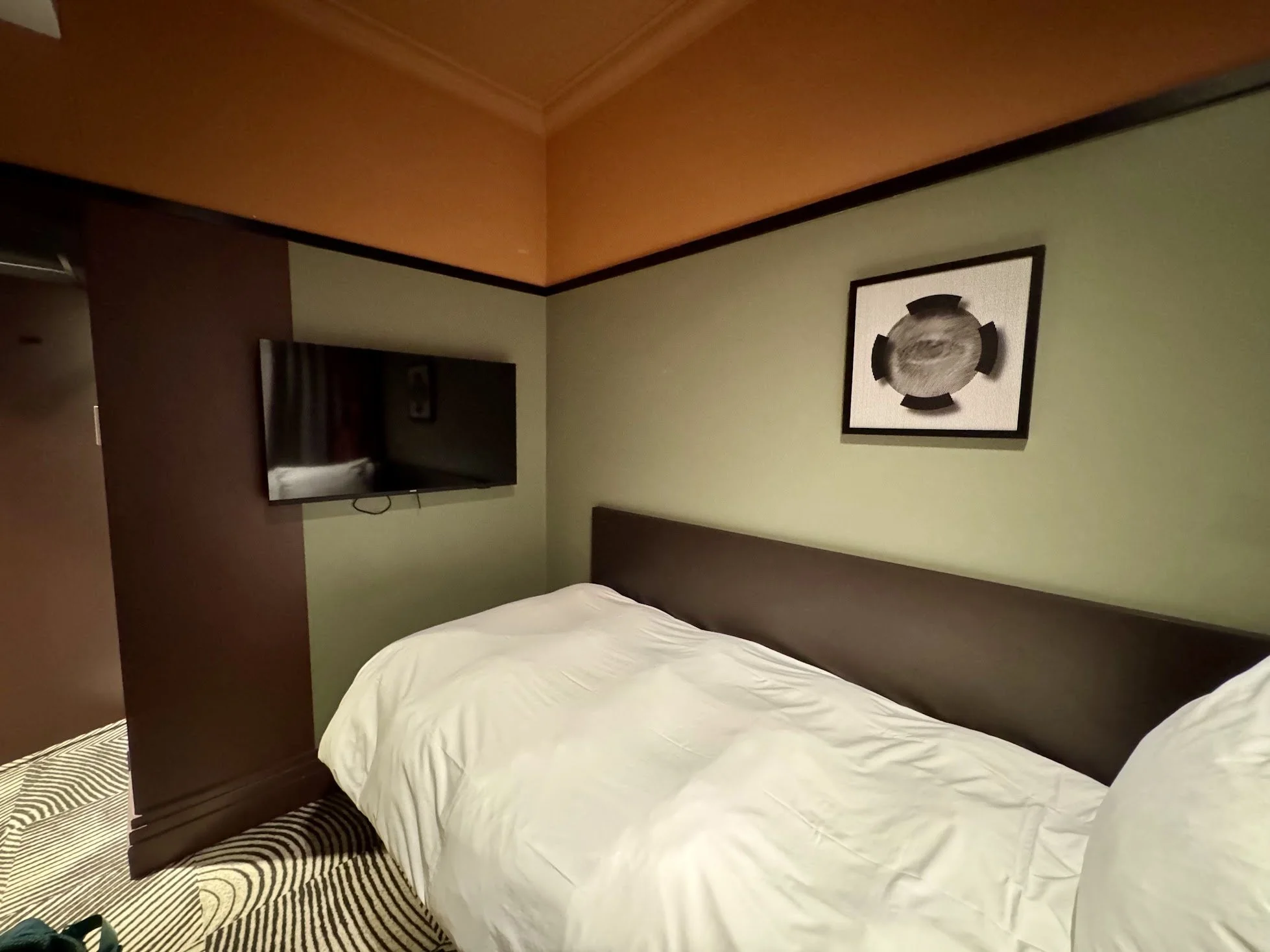 A hotel room with a bed, a wall-mounted television, and framed artwork. The room has a green wall, a brown accent wall, and an orange ceiling, with striped carpet.